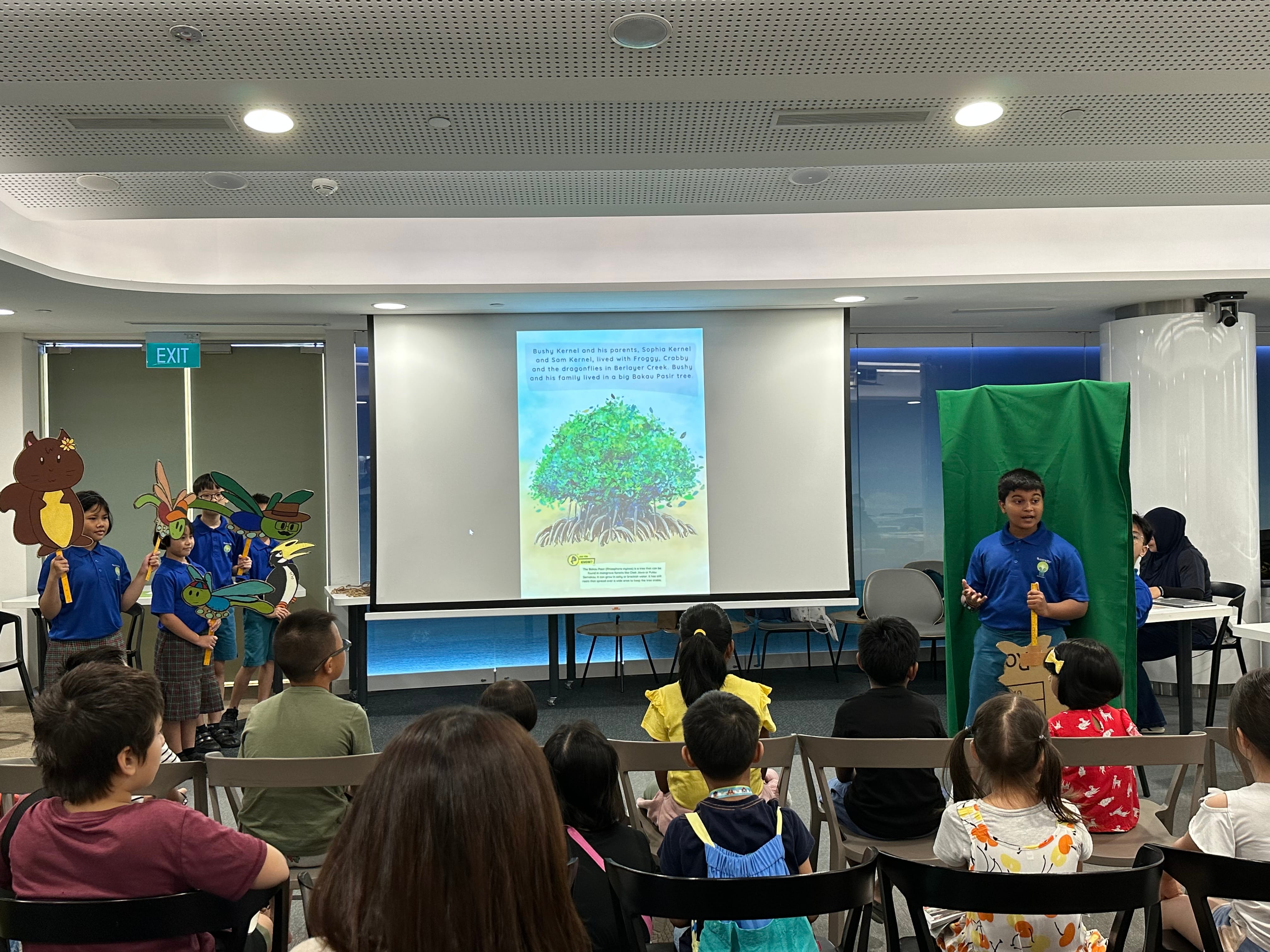 Children in blue shirts perform a puppet show for a seated audience, with a projected storybook tree slide behind them.