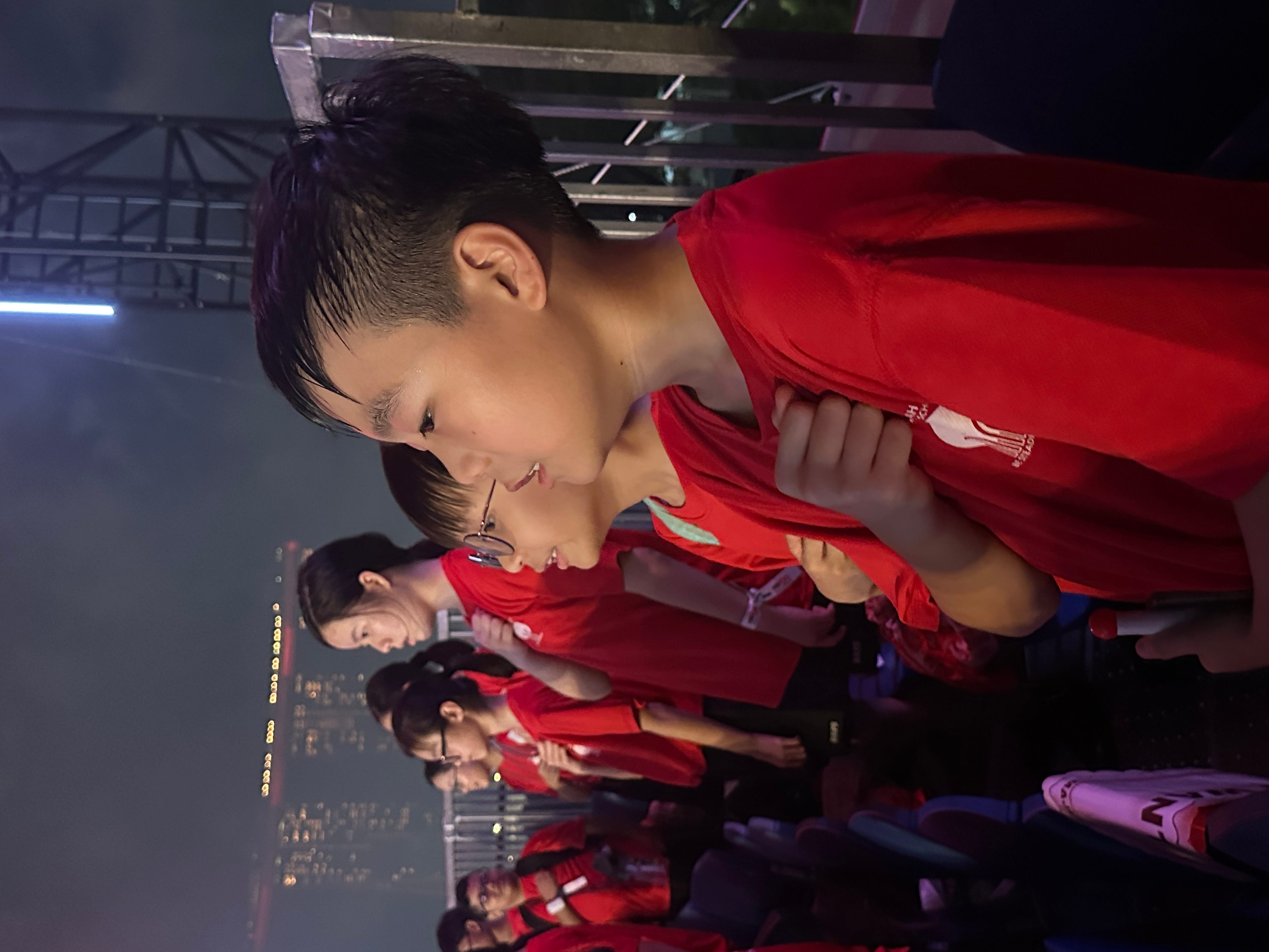 A row of children in red shirts stand with hands on chests at an outdoor night event with city lights behind them.