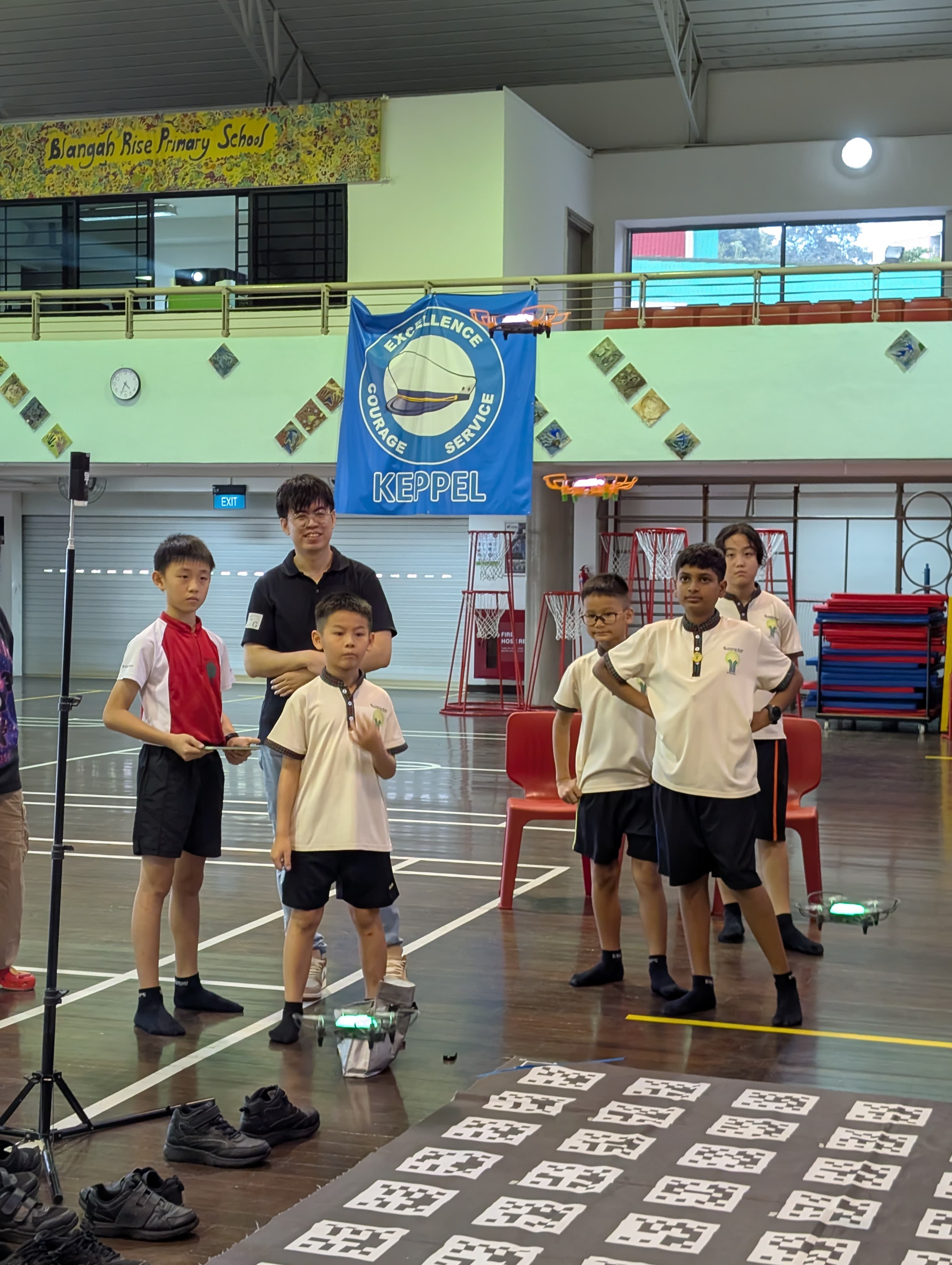 Students and a teacher stand in a school gym at Blangah Rise Primary School during a drone flying activity.