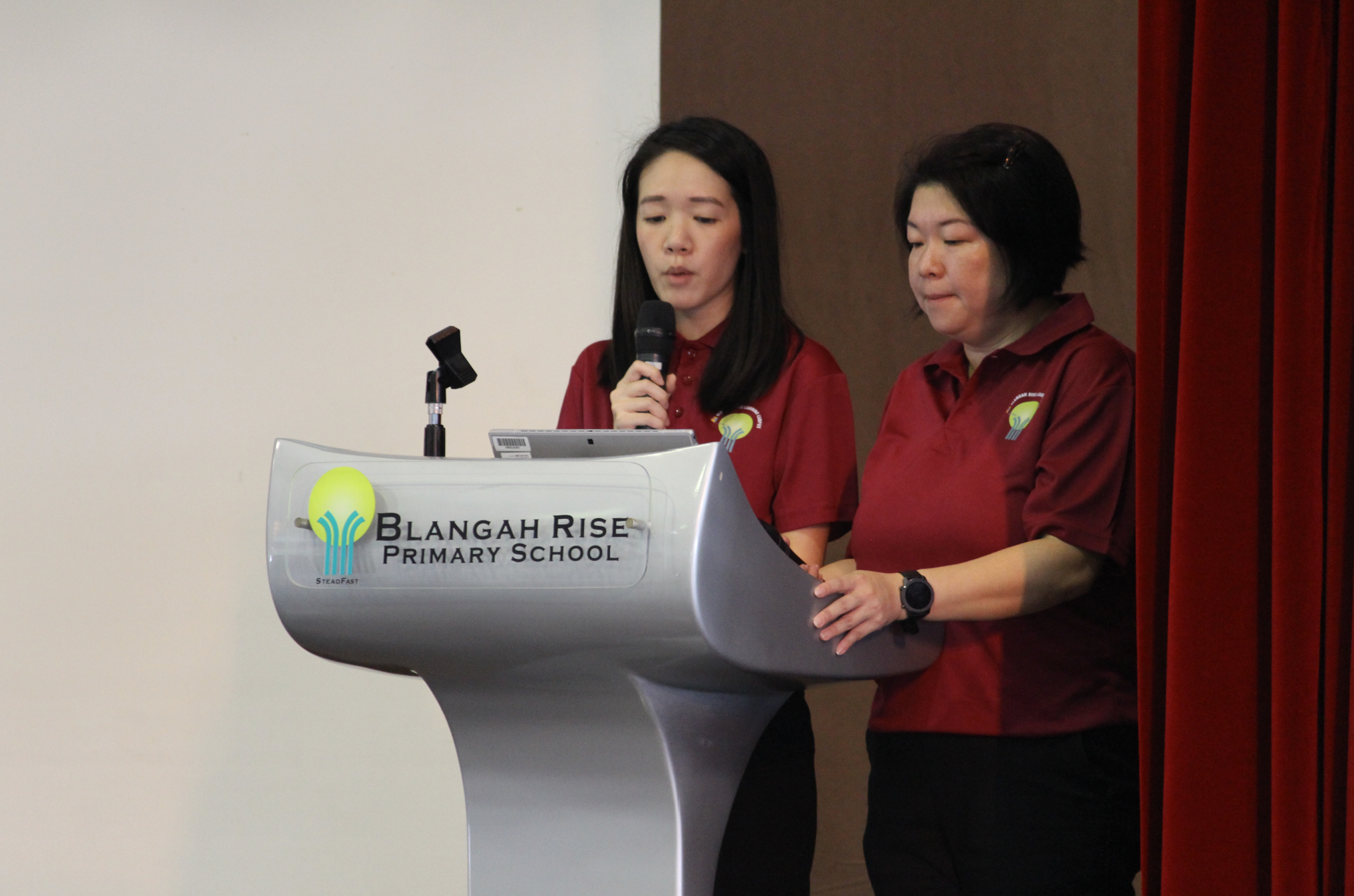 Two women in red shirts stand at a Blangah Rise Primary School podium, one speaking into a microphone.