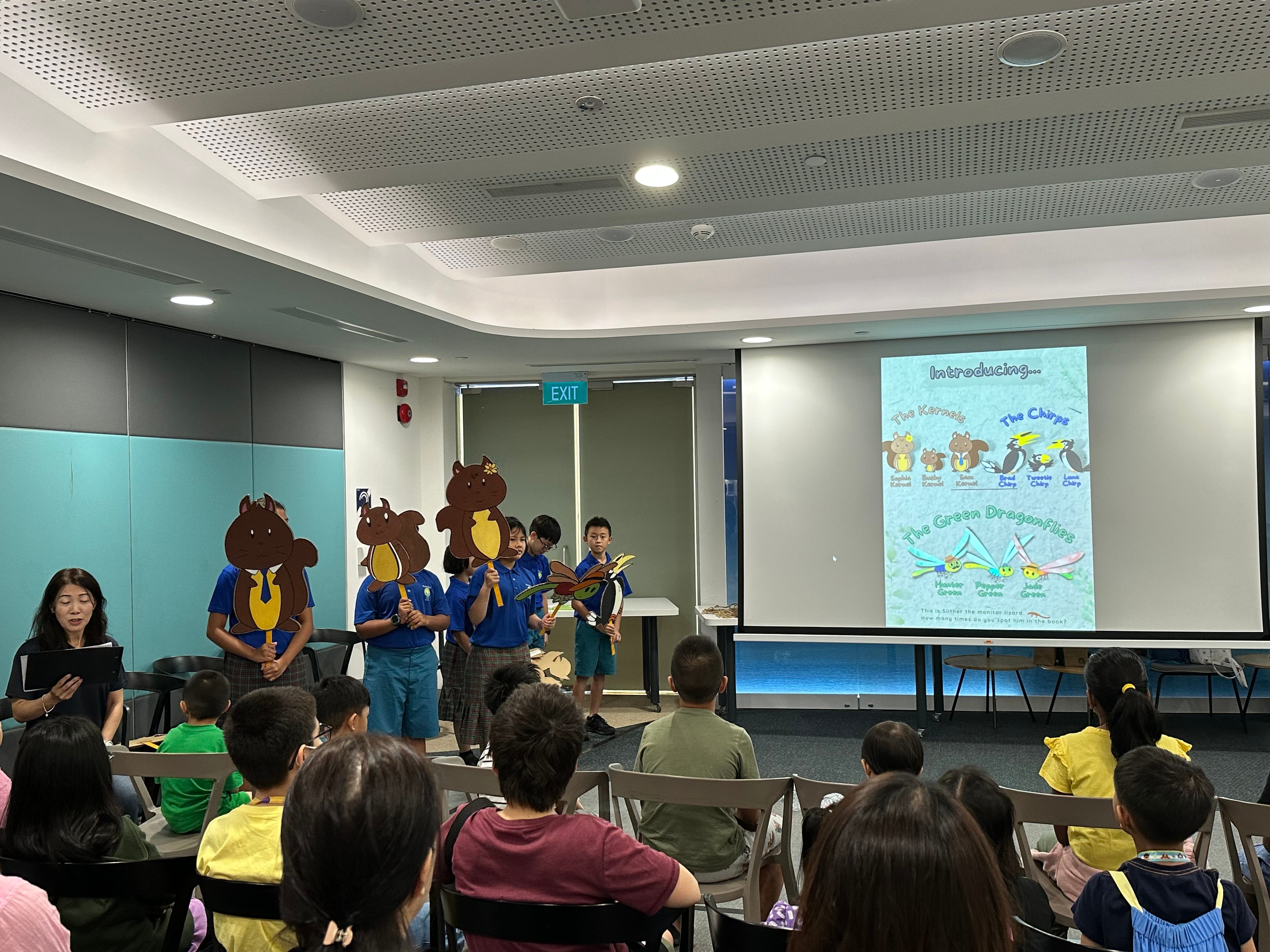 Children in blue shirts hold animal cutouts while presenting to a seated audience, with a cartoon character slideshow projected behind them.