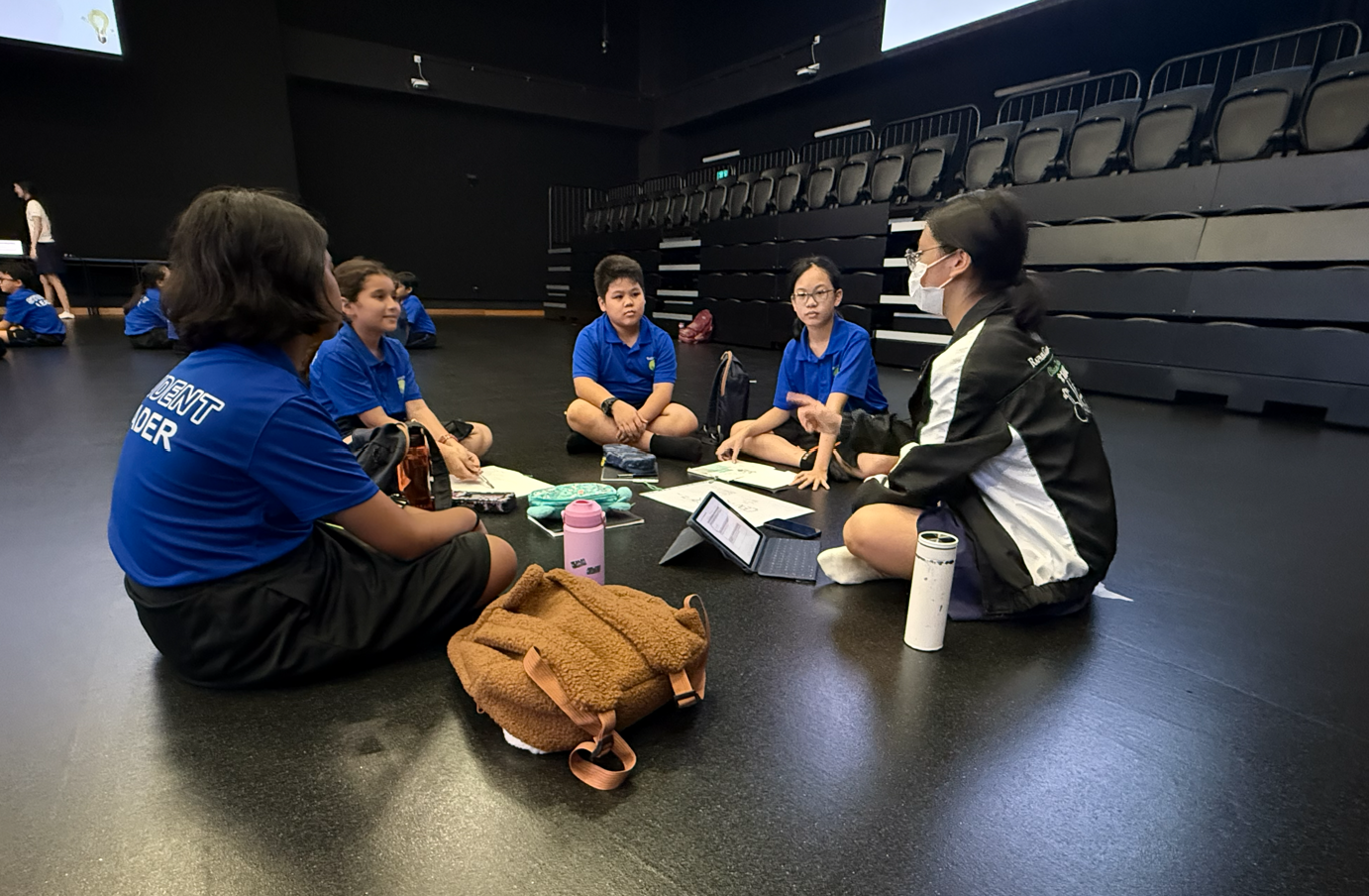 Group in blue shirts sits on floor of dark auditorium, discussing papers and a laptop with water bottles nearby
