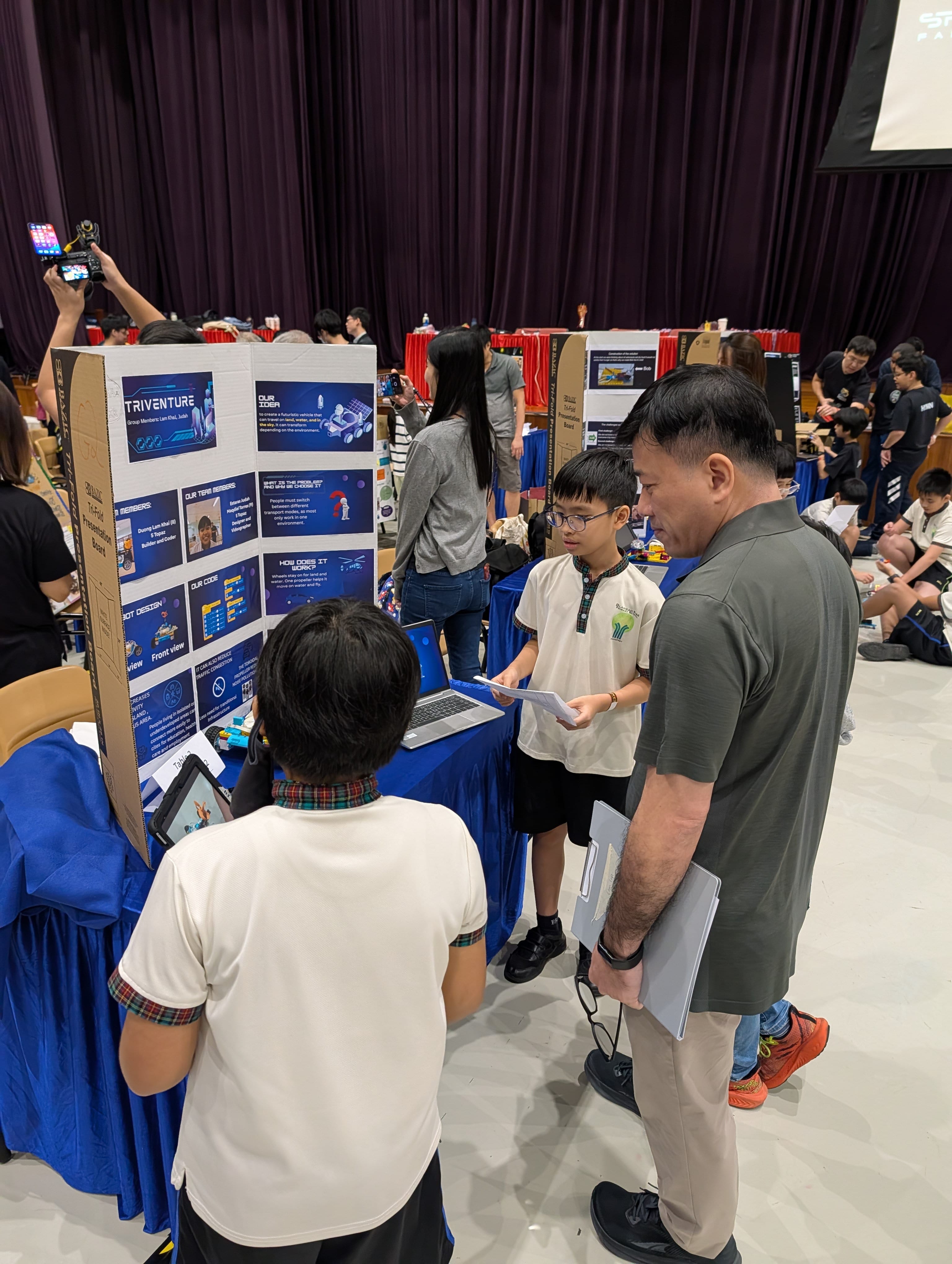 A student presents a science fair project board labeled Triventure to an adult judge in a busy school hall.