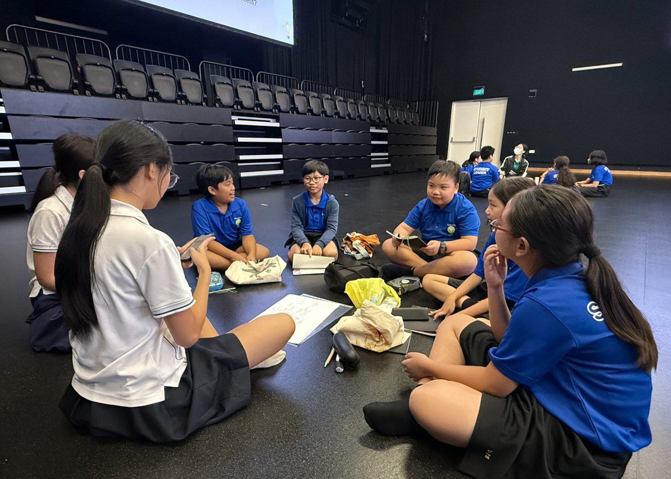 Six students in blue uniforms sit in a circle on a dark floor in an auditorium, engaged in group discussion.