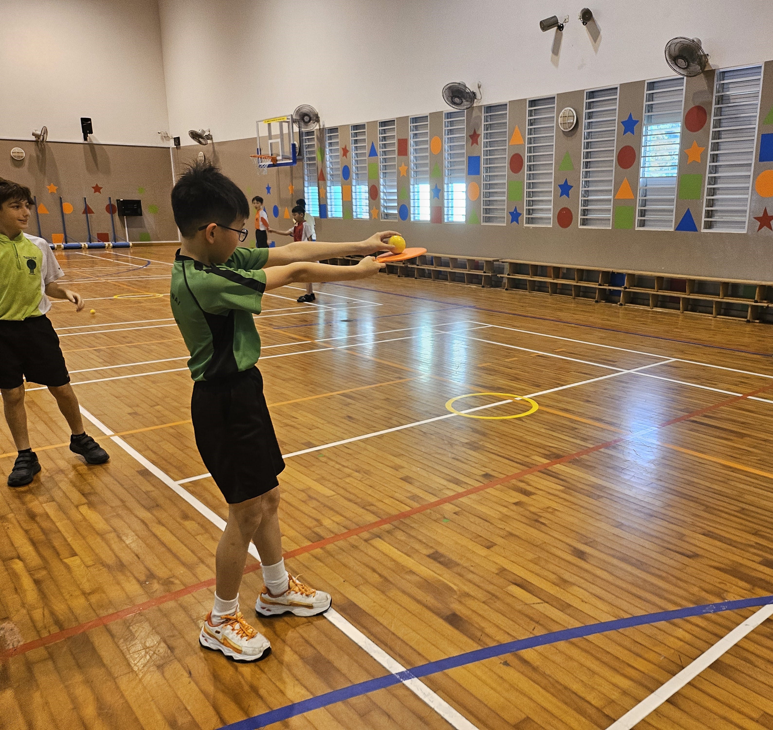 A boy in a green shirt holds an orange paddle with a yellow ball in a colorful school gymnasium.