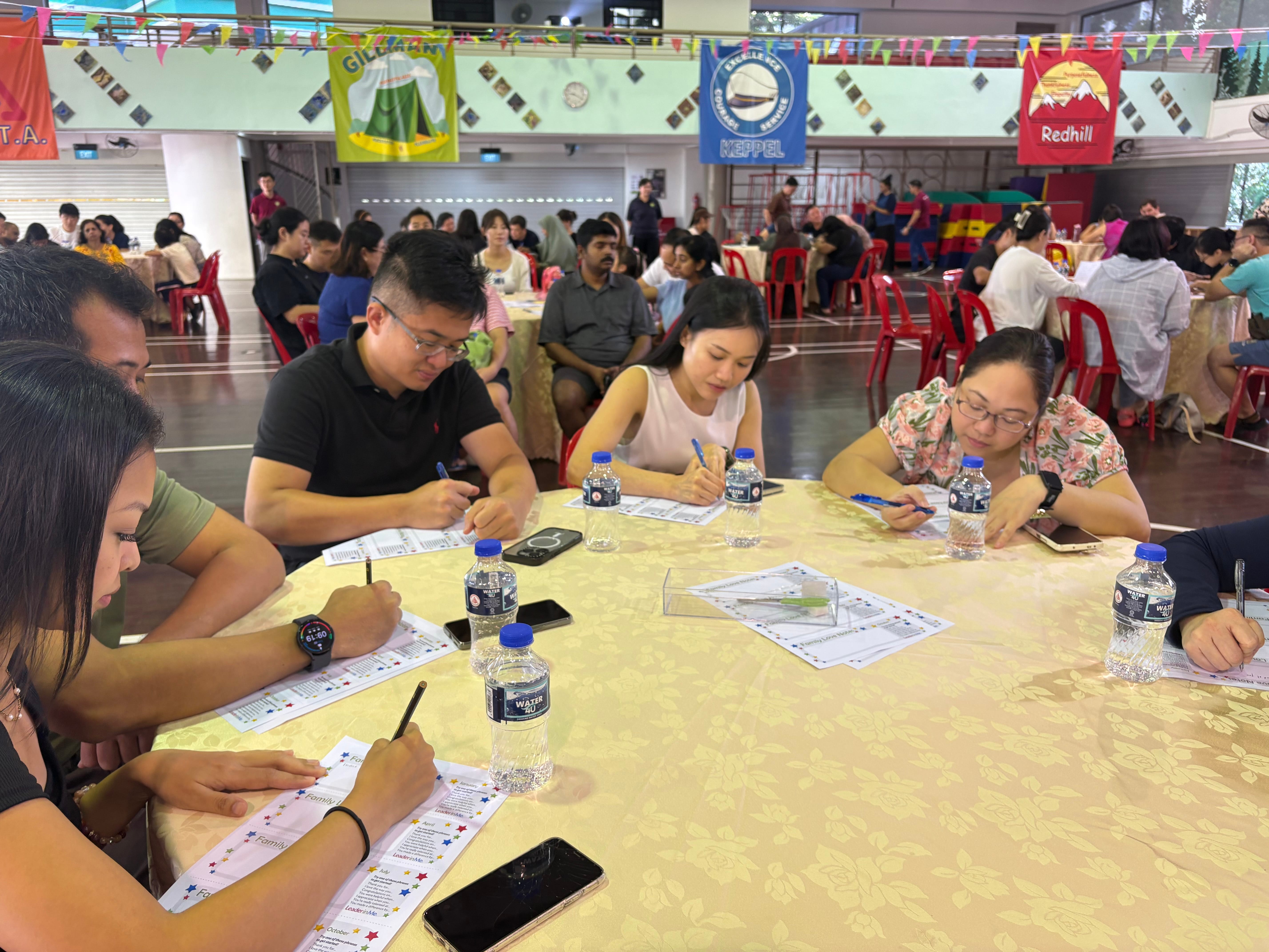 People seated at round tables in a hall, writing on worksheets, with water bottles and phones on table