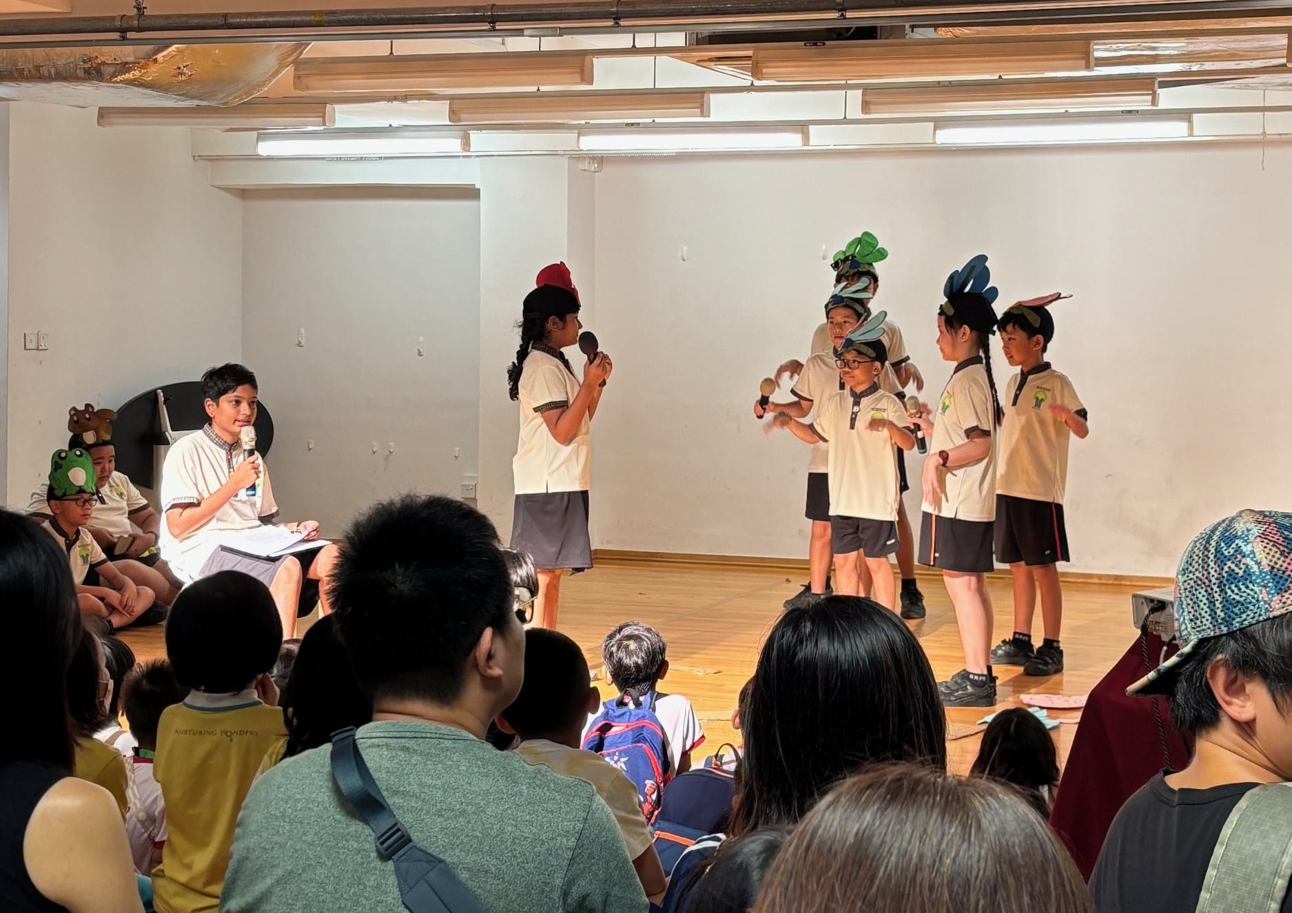 Children in school uniforms perform a skit on stage wearing costume hats, holding microphones, before a seated audience.