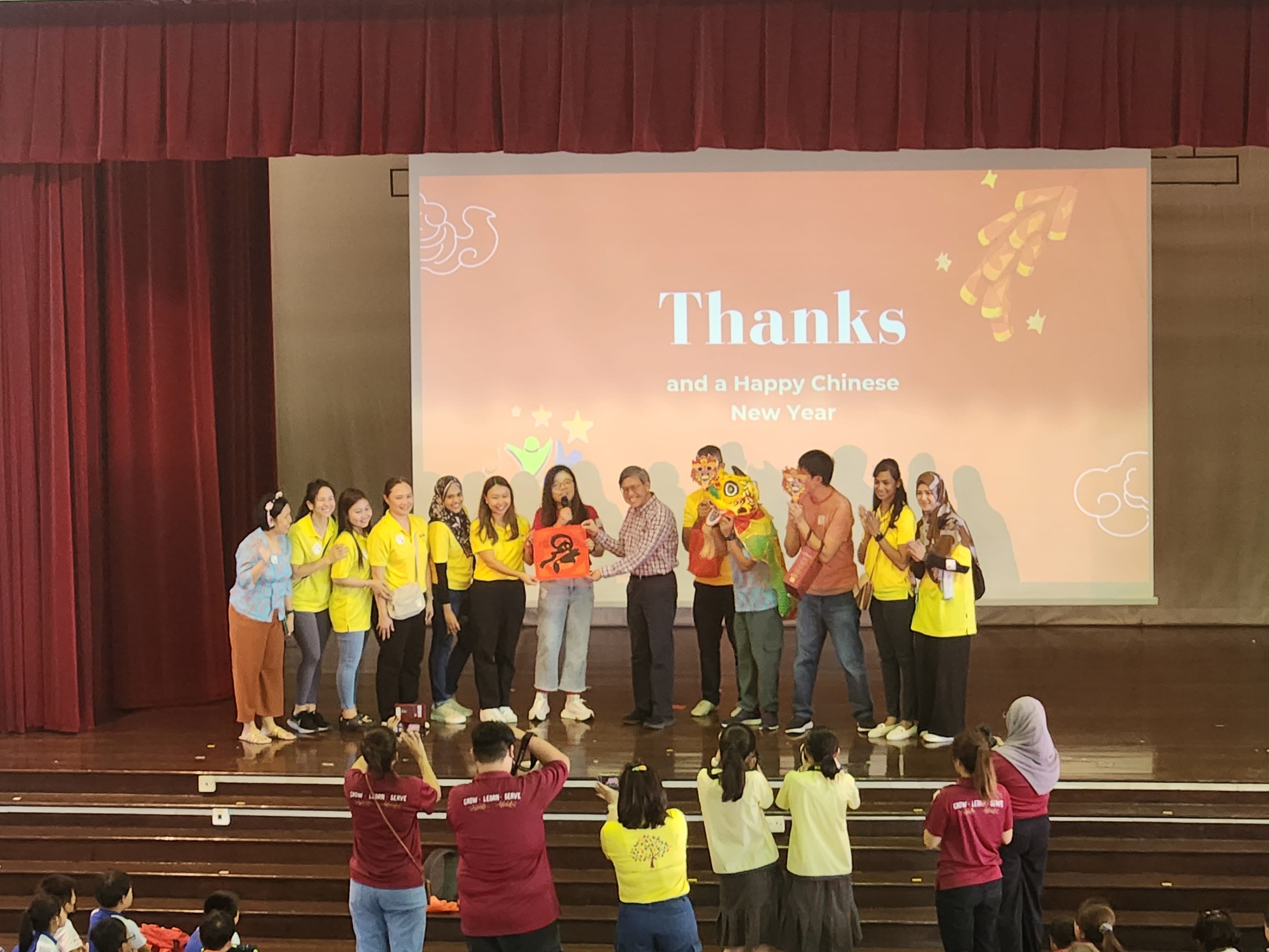 Group of people on stage at a Chinese New Year event, with a projected screen reading "Thanks and a Happy Chinese New Year."