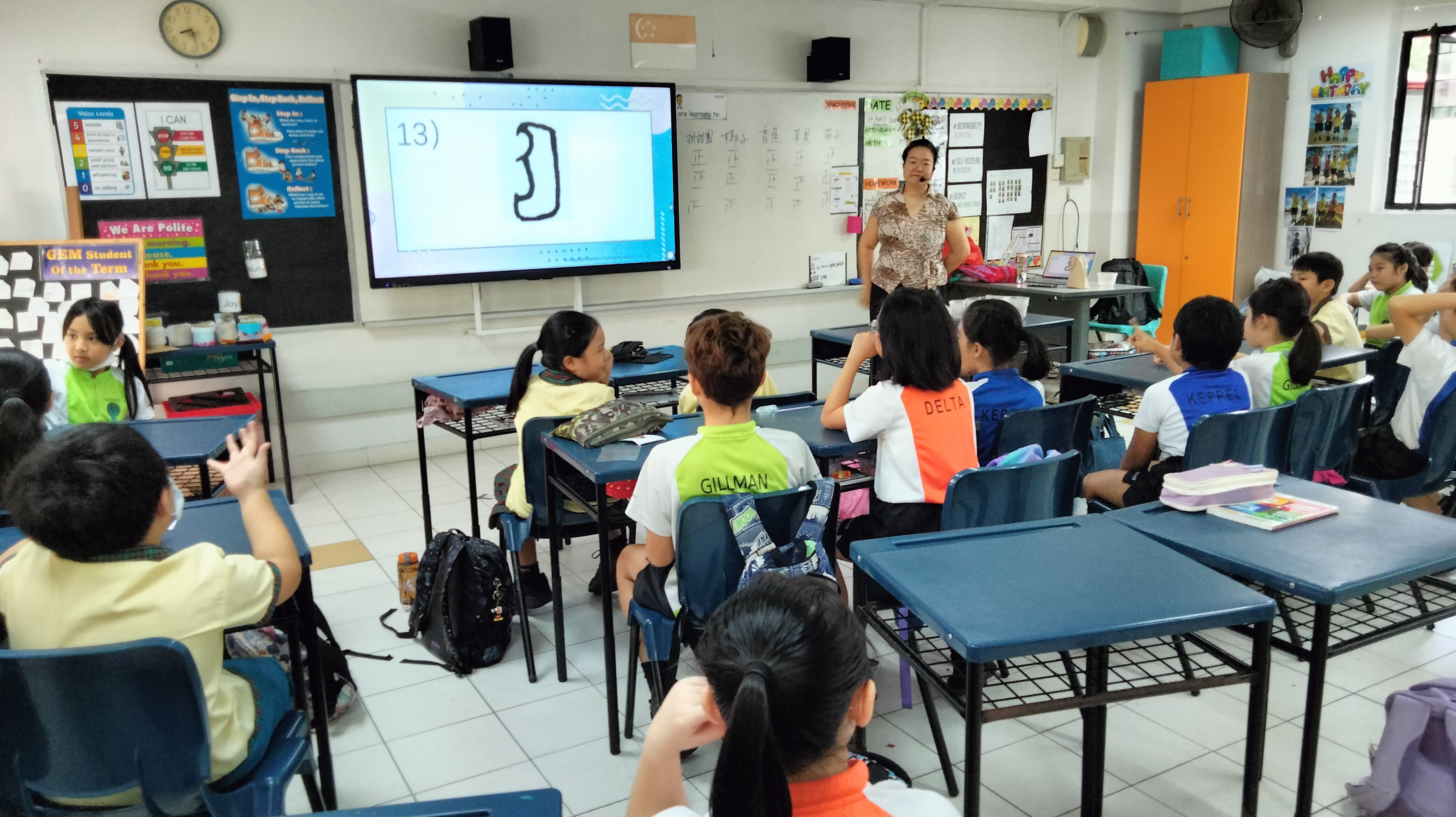 A teacher stands at the front of a classroom with students at desks facing a large screen showing number 13.