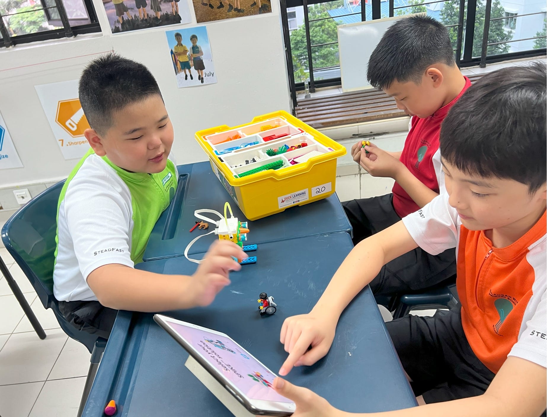 Three boys sit at a desk building with LEGO pieces, using a tablet and a yellow parts storage bin.
