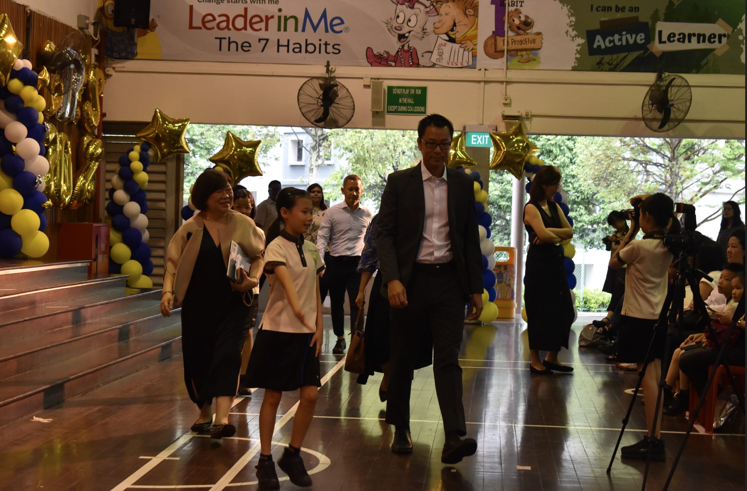 Adults and a child walk through a school hall decorated with balloons under a Leader in Me 7 Habits banner.