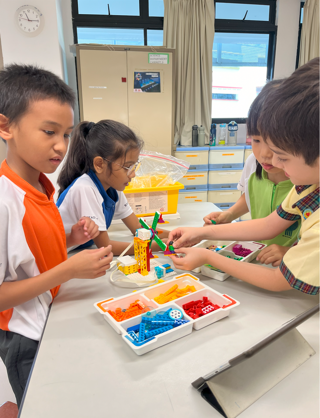 Four children work together at a table building a colorful LEGO model using sorted brick trays in a classroom.
