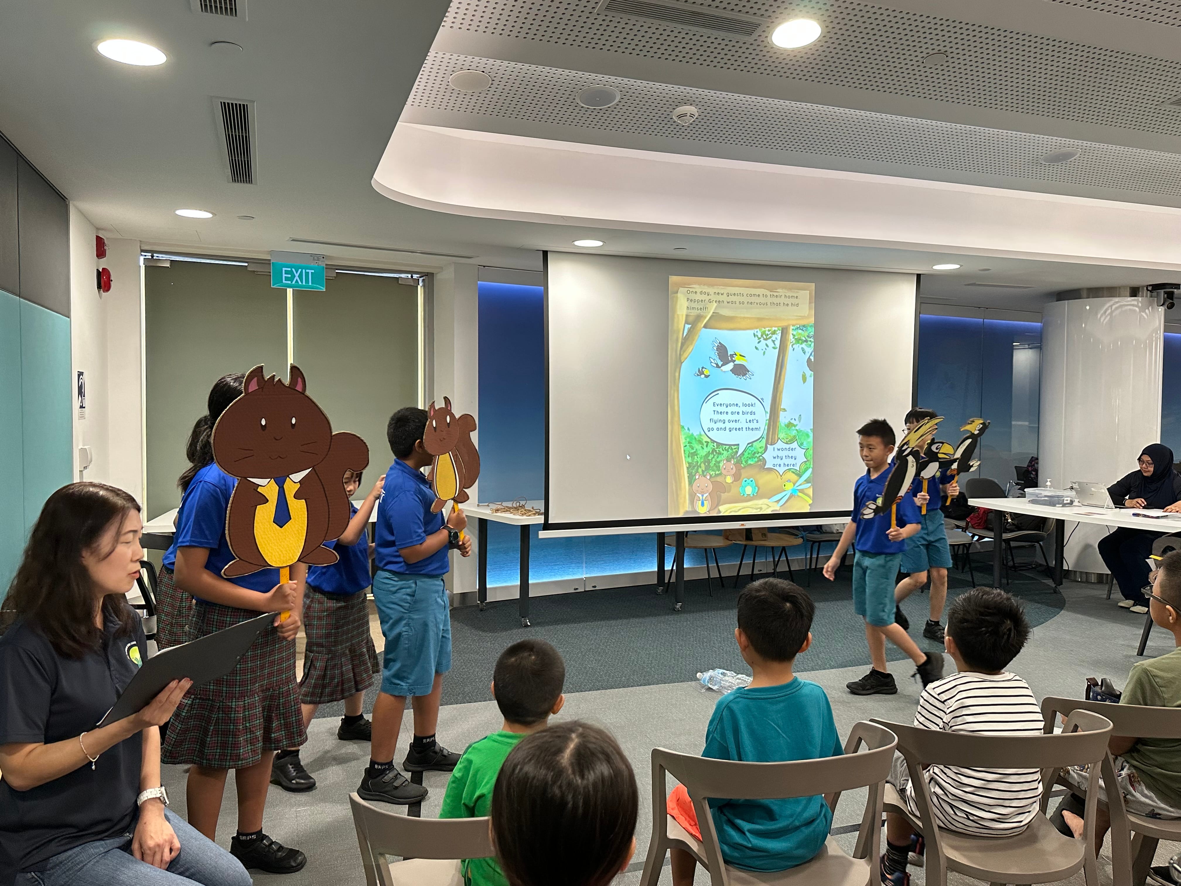 Children in blue uniforms hold large animal cutout puppets during a storytime performance in front of a projected storybook.