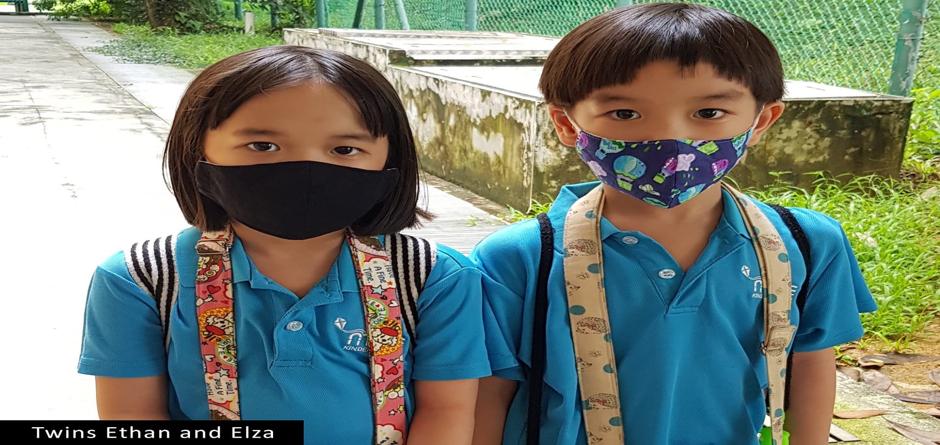 Two young children in blue school uniforms and face masks stand together outdoors wearing backpacks.