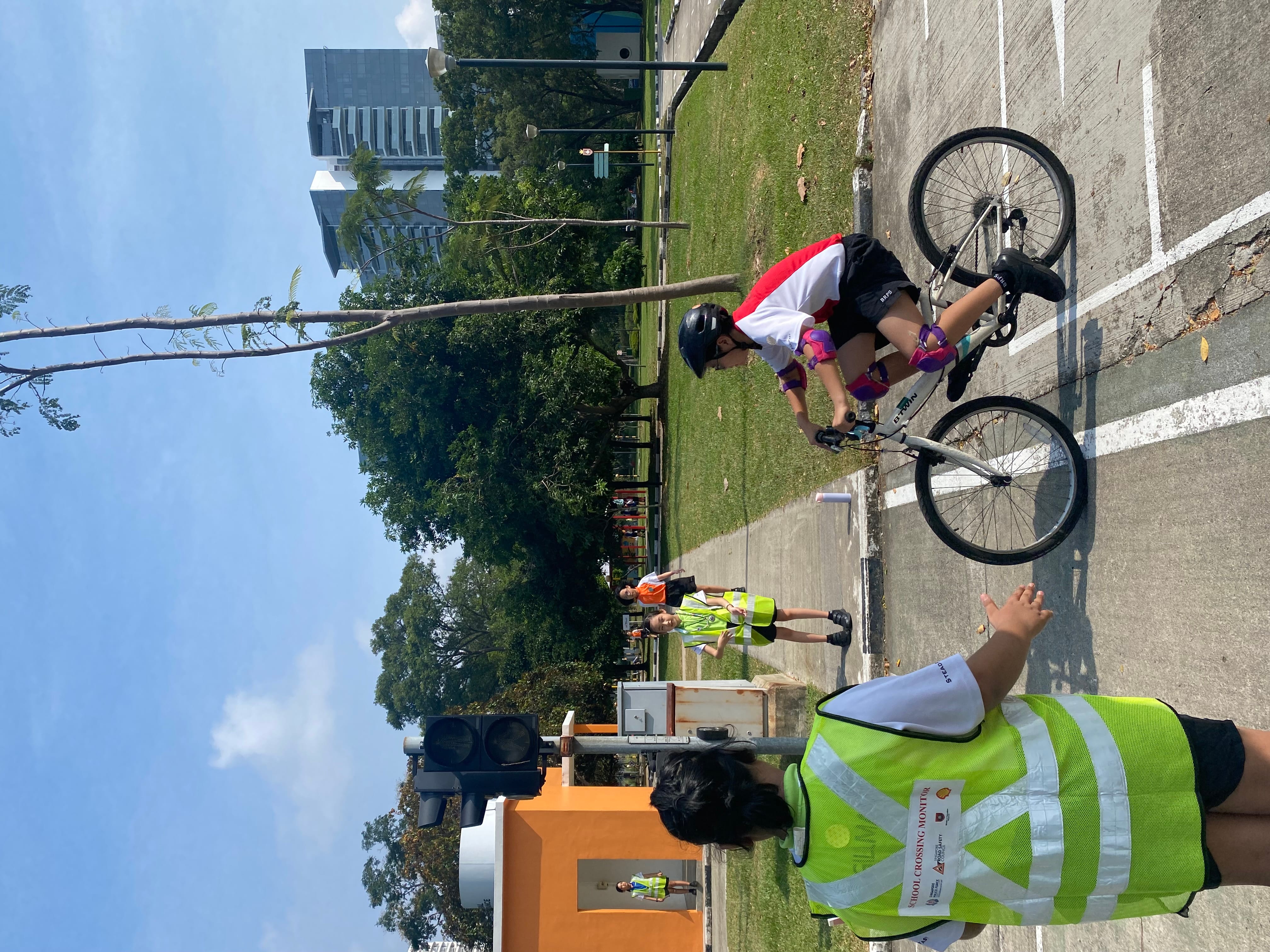 A child on a bicycle approaches a crosswalk while a school crossing monitor in a yellow vest signals to stop.