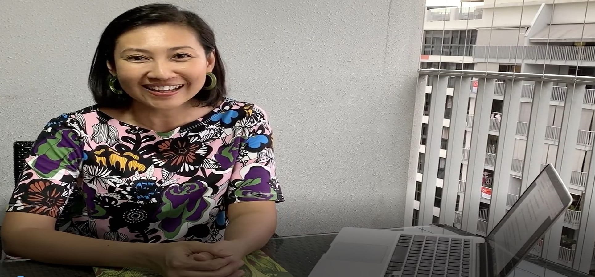 Smiling woman in a colorful floral top sits at a table with a laptop, with a city building visible behind her.