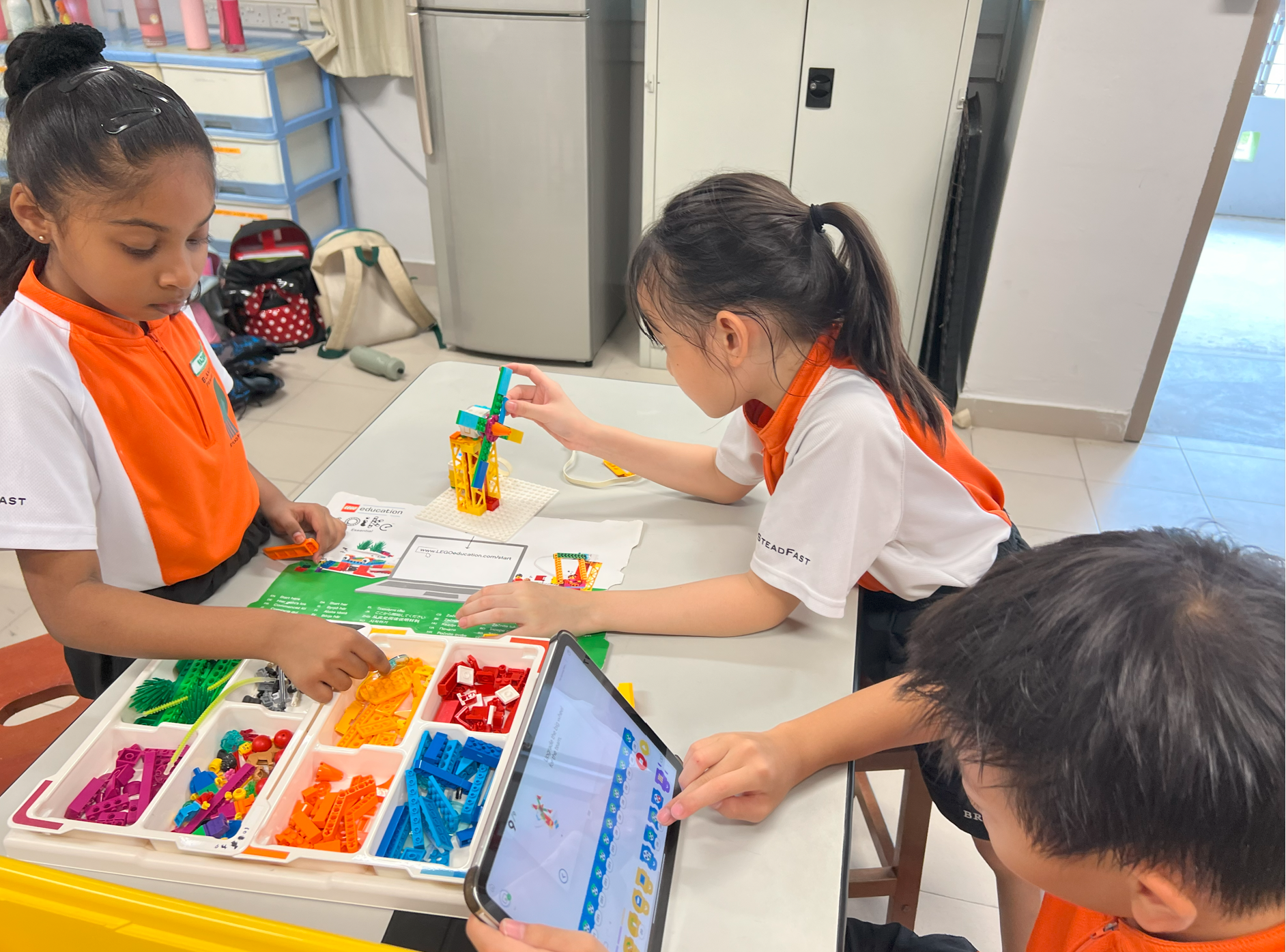 Three children in orange-and-white uniforms build a LEGO model at a table, one using a tablet.
