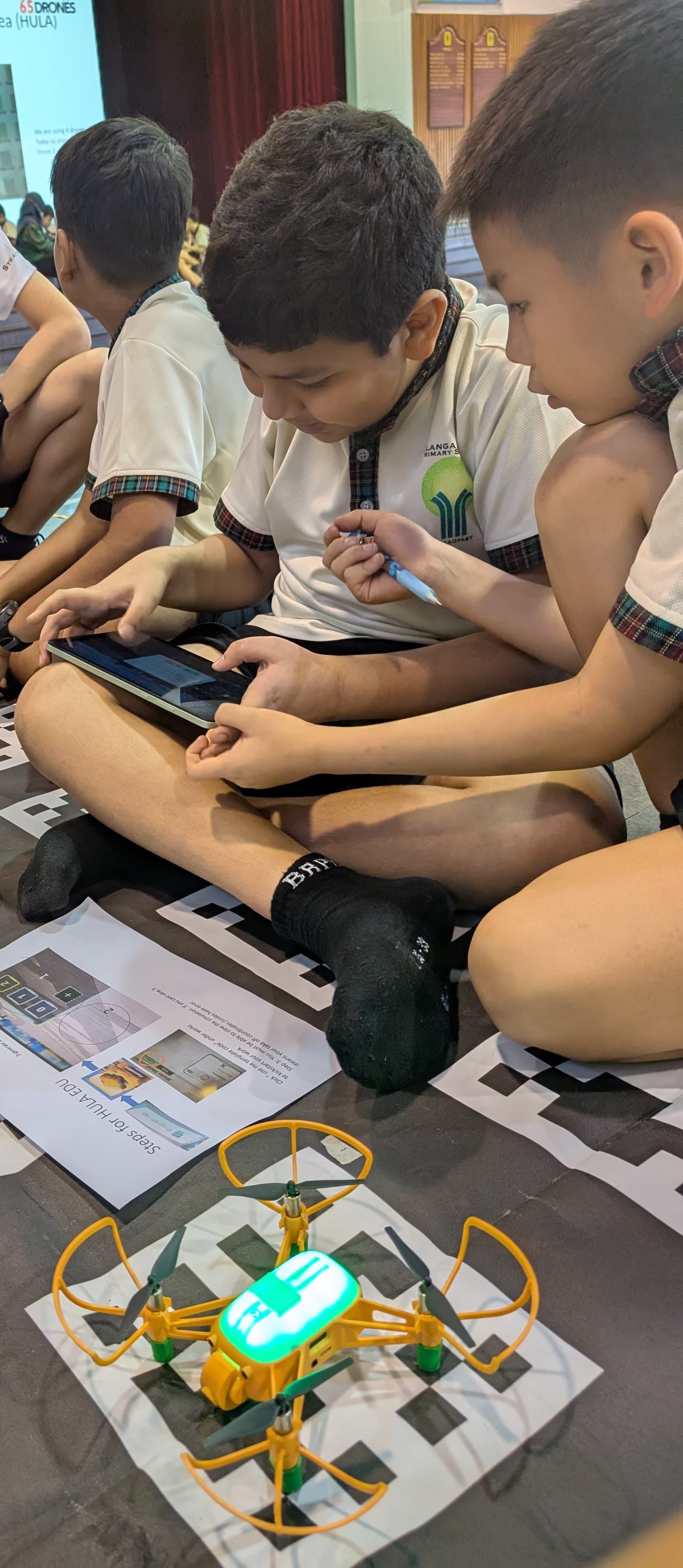 Two school children sit on the floor using a tablet to control a small yellow drone with green lights.