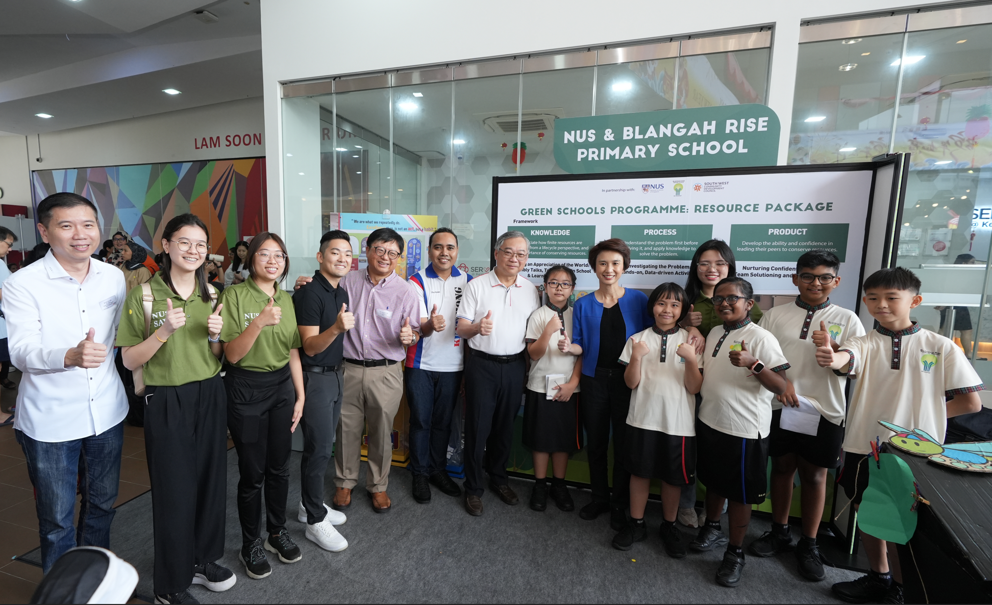 Group of adults and schoolchildren giving thumbs up in front of a “NUS & Blangah Rise Primary School” display.