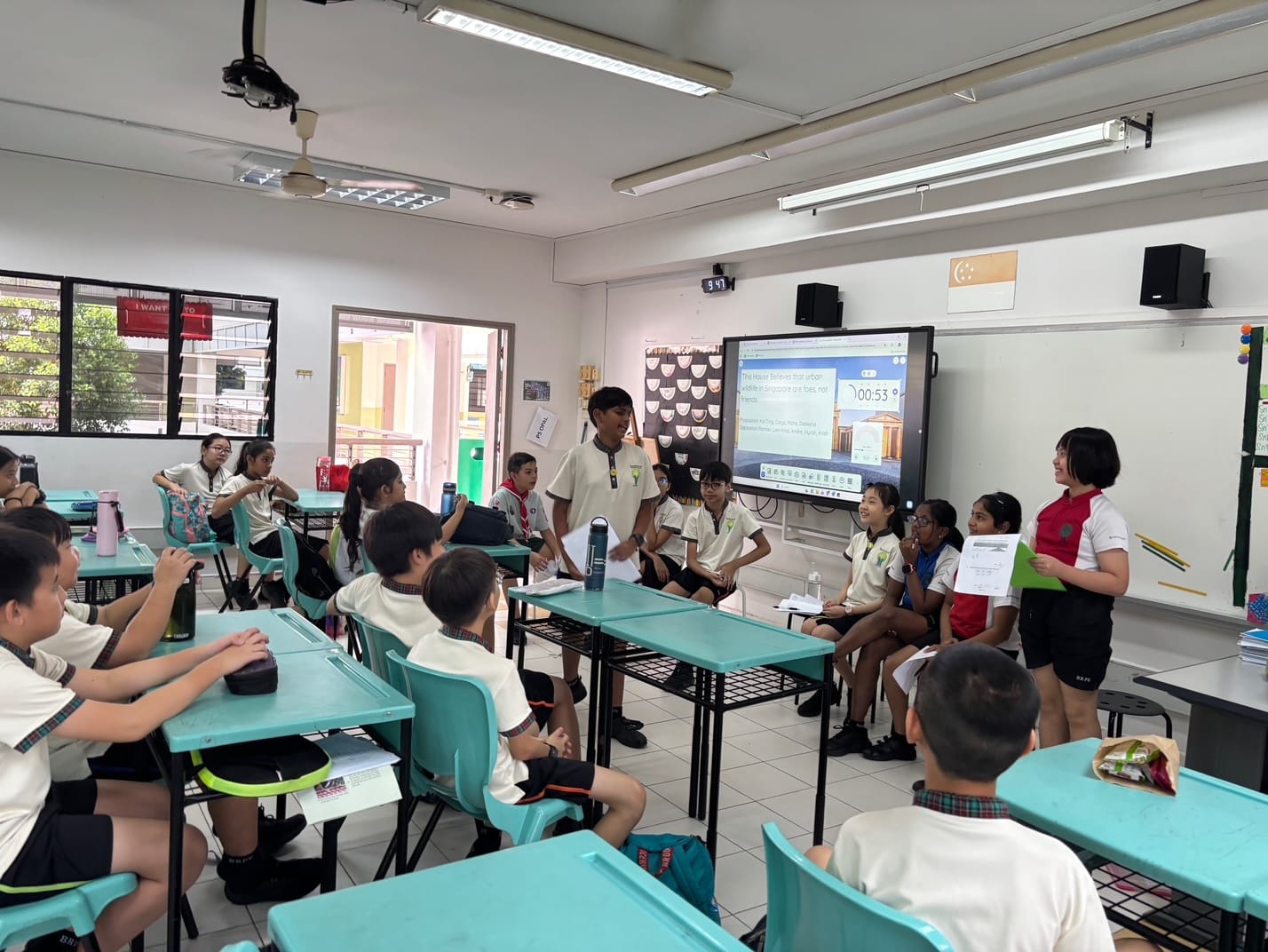 Students in uniforms sit at teal desks in a classroom while two pupils stand presenting near a screen showing a timer.