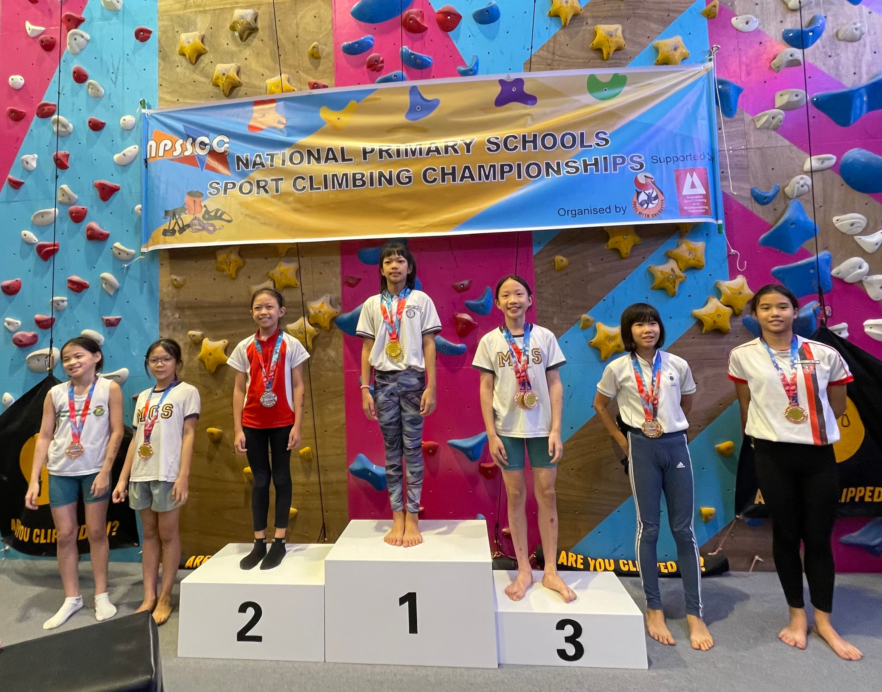 Six girls with medals stand on podiums at the National Primary Schools Sport Climbing Championships in front of a colorful climbing wall.