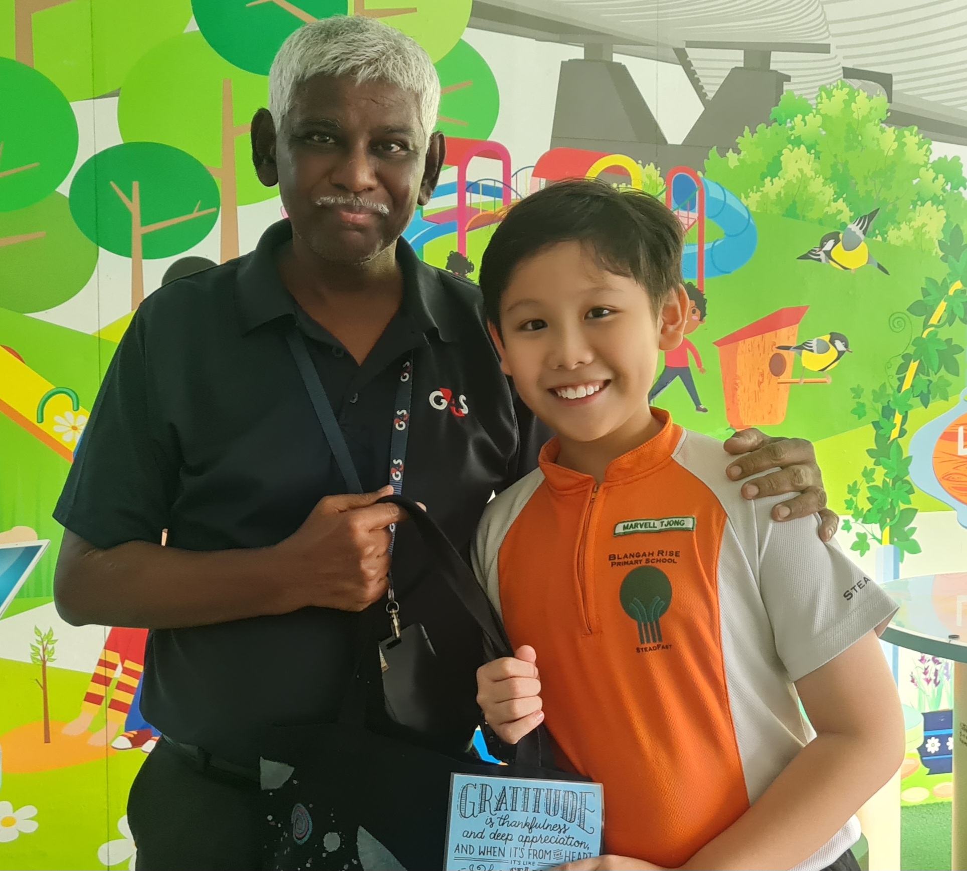 An elderly man with white hair and a G4S uniform poses with a smiling boy in an orange school uniform.