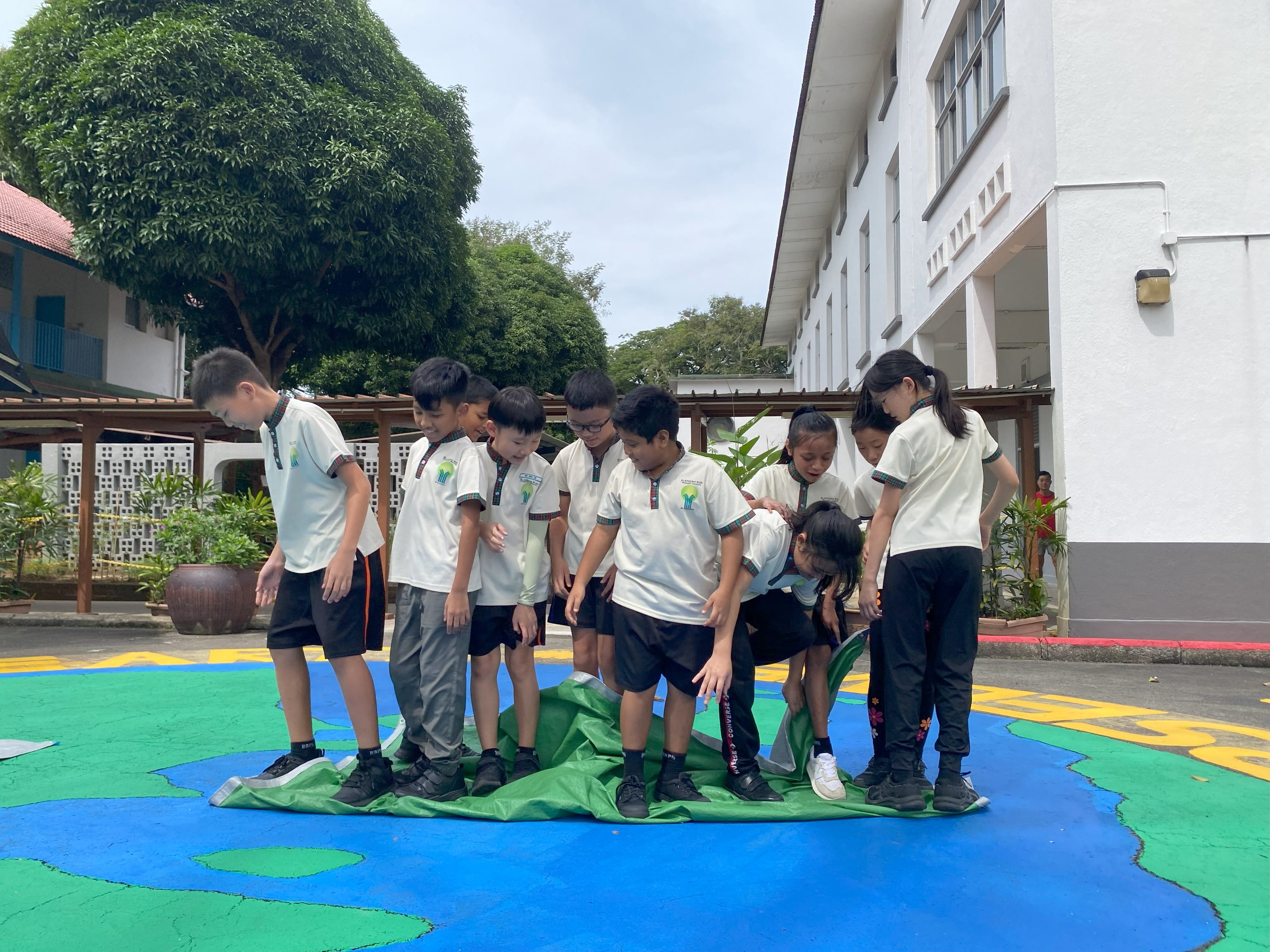 A group of children in white school uniforms stand on a colorful painted ground mural outdoors near a white building.