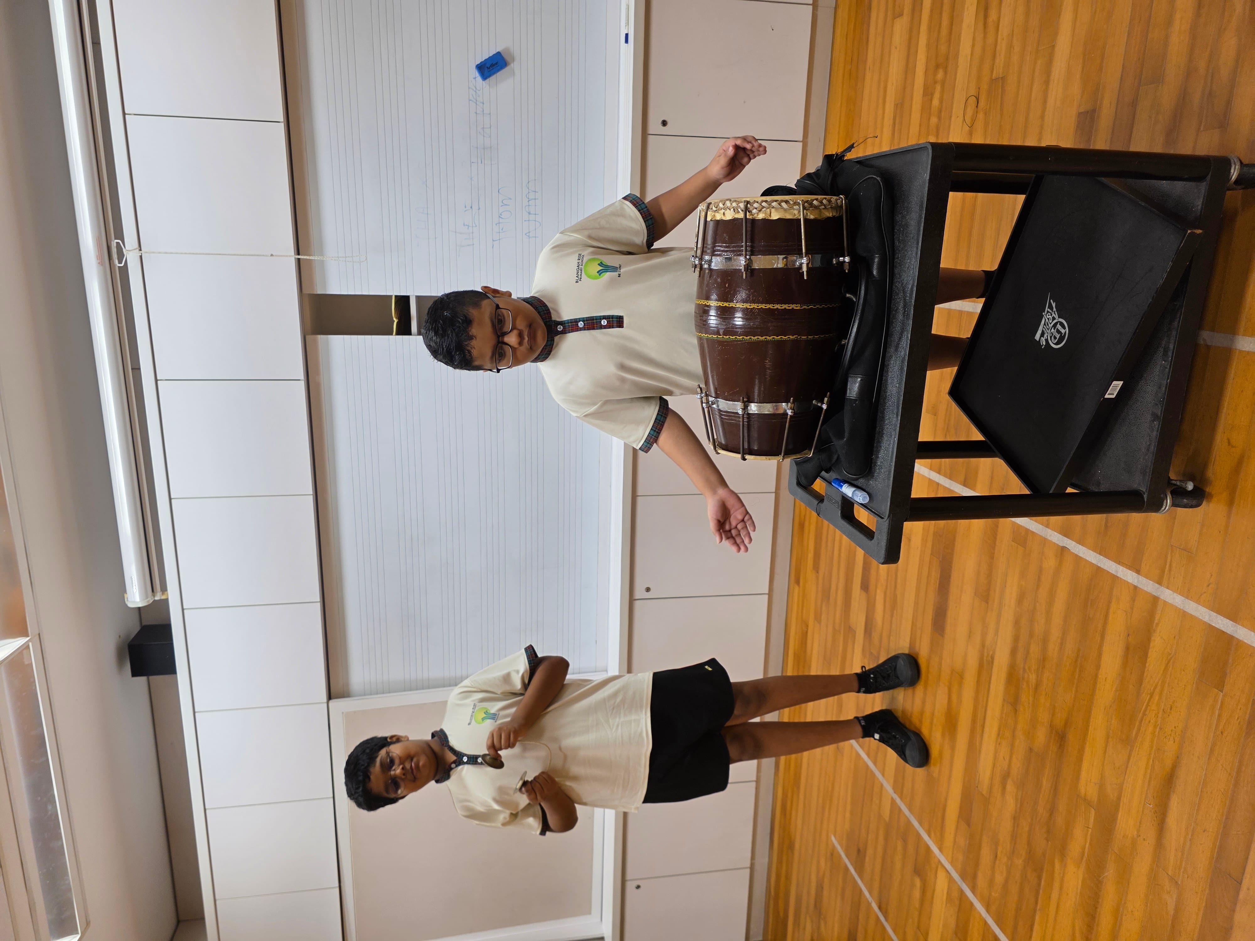 Two boys in school uniforms in a classroom, one playing a dholak drum on a cart, the other holding sticks.