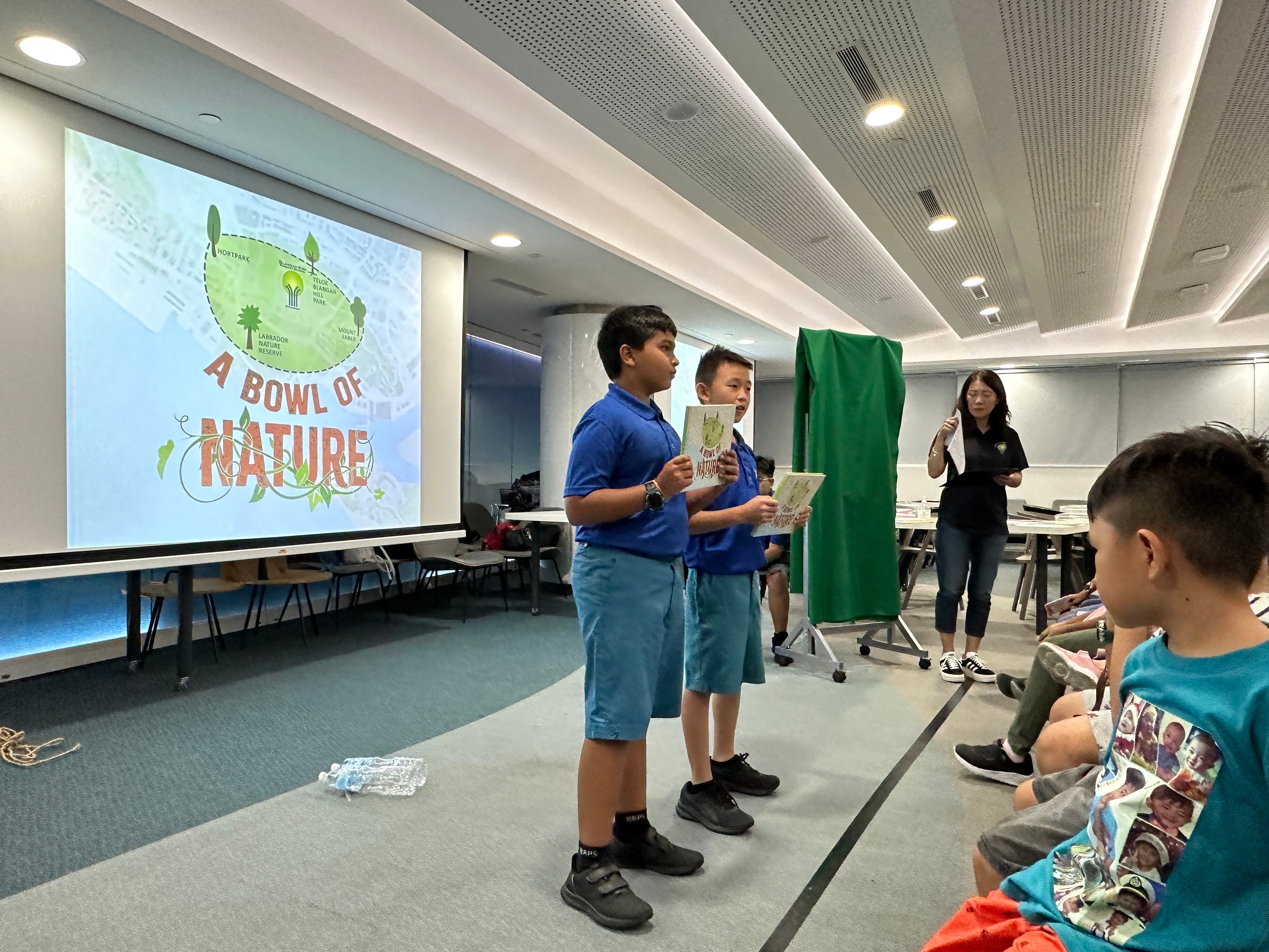 Two boys in blue uniforms present in front of a projected slide reading "A Bowl of Nature" to classmates.