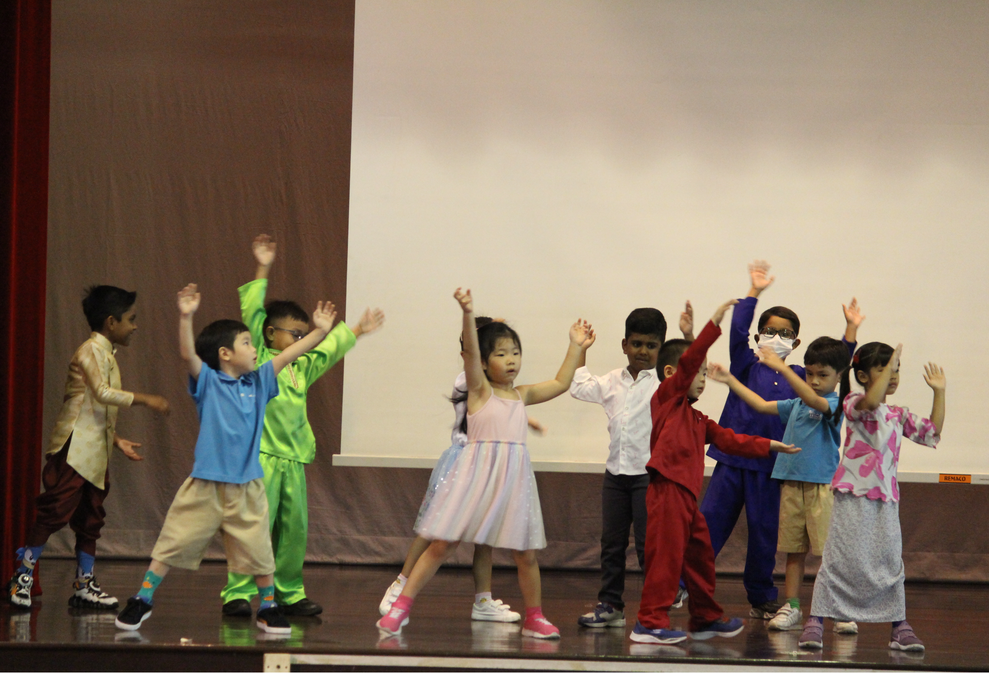 Group of children on a stage dancing with arms raised, wearing colorful outfits and costumes.