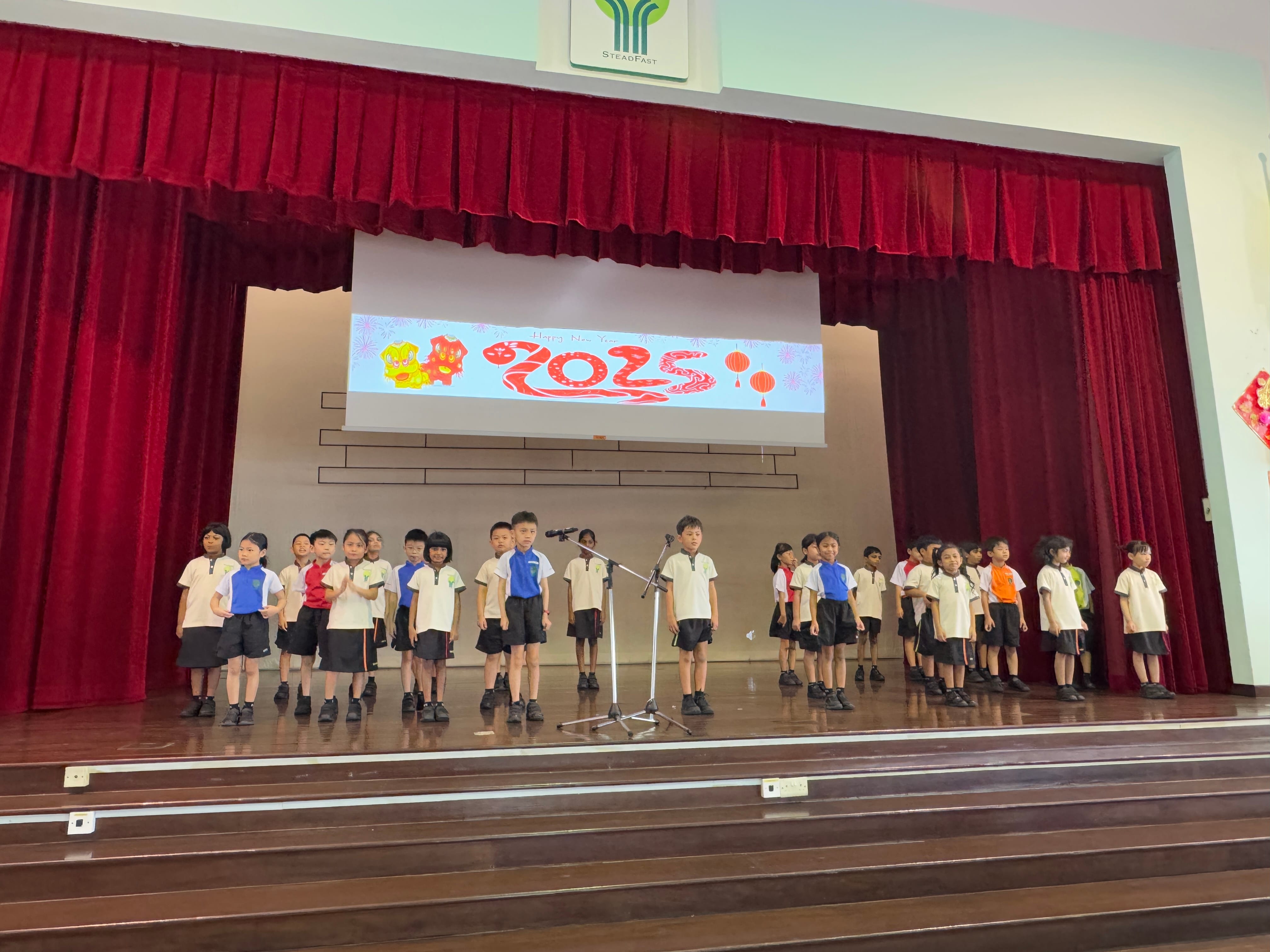 Young students in school uniforms stand on a stage with red curtains and a 2025 Chinese New Year backdrop.