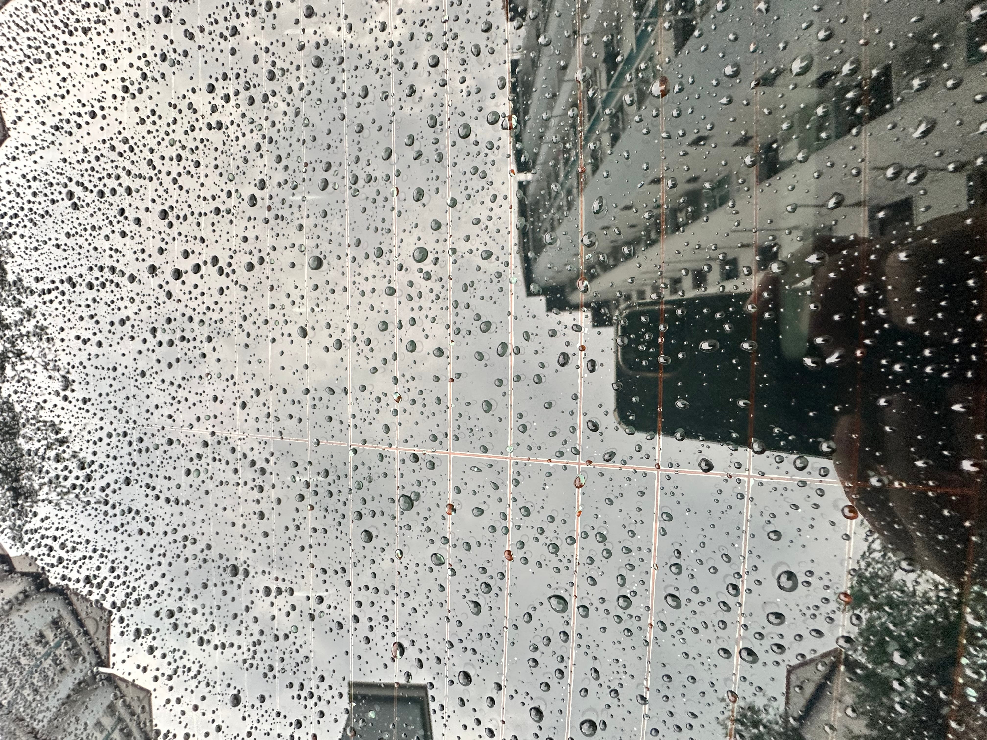 Raindrops on a glass panel with grid lines, reflecting a building and a hand holding a phone.
