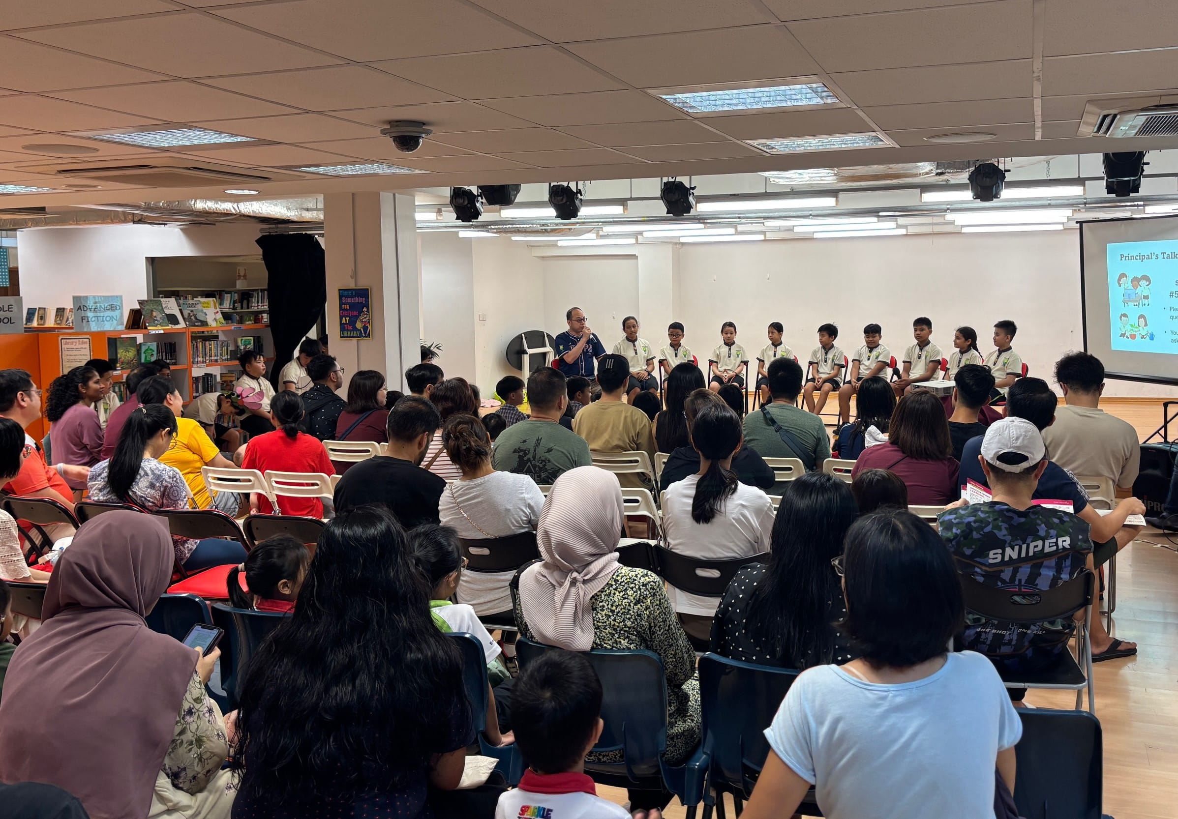 A crowded school hall with seated adults facing a row of students on stage during a Principal's Talk event.