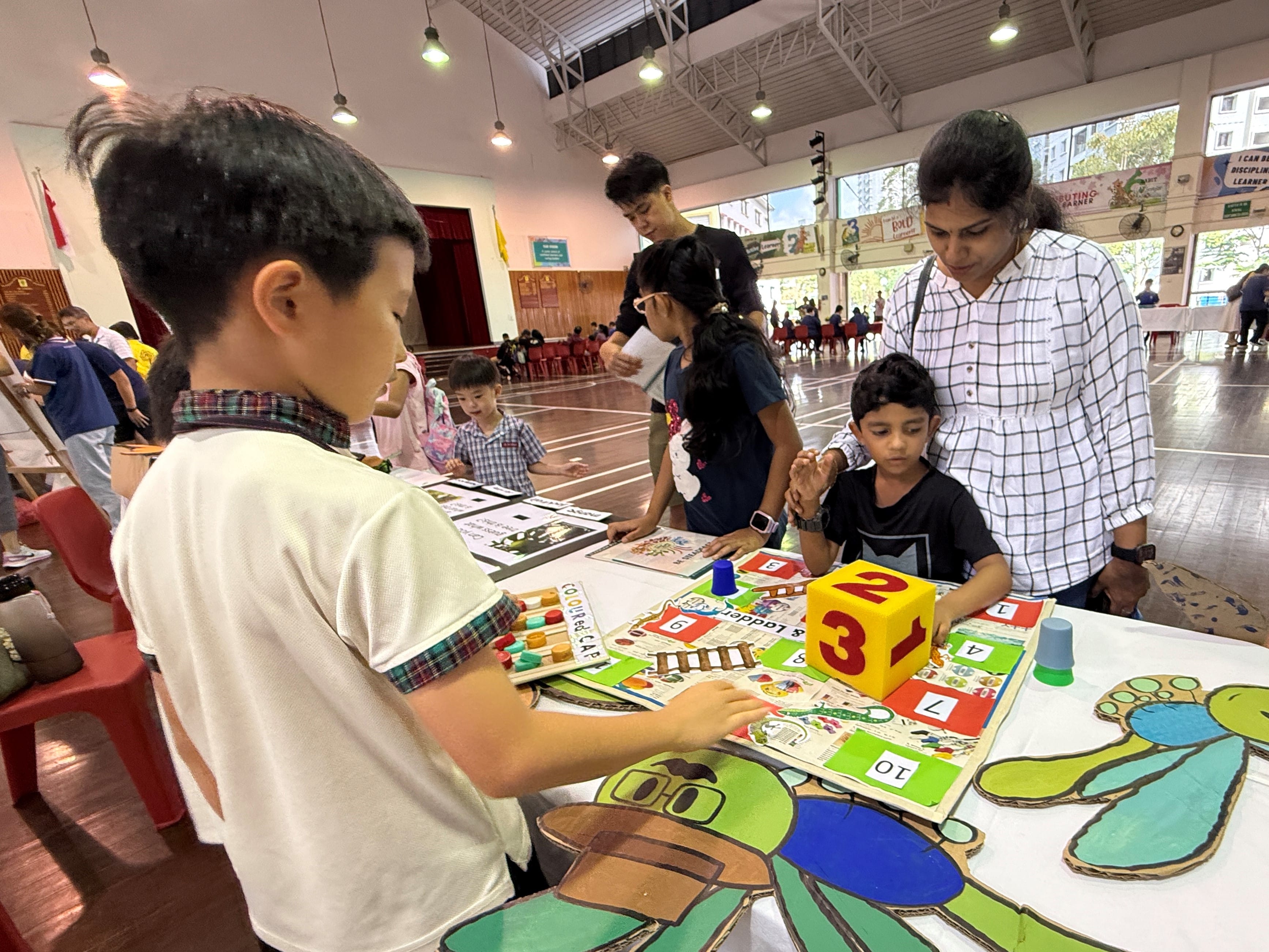 Children and an adult play a tabletop number board game with a large foam die in a bright hall.