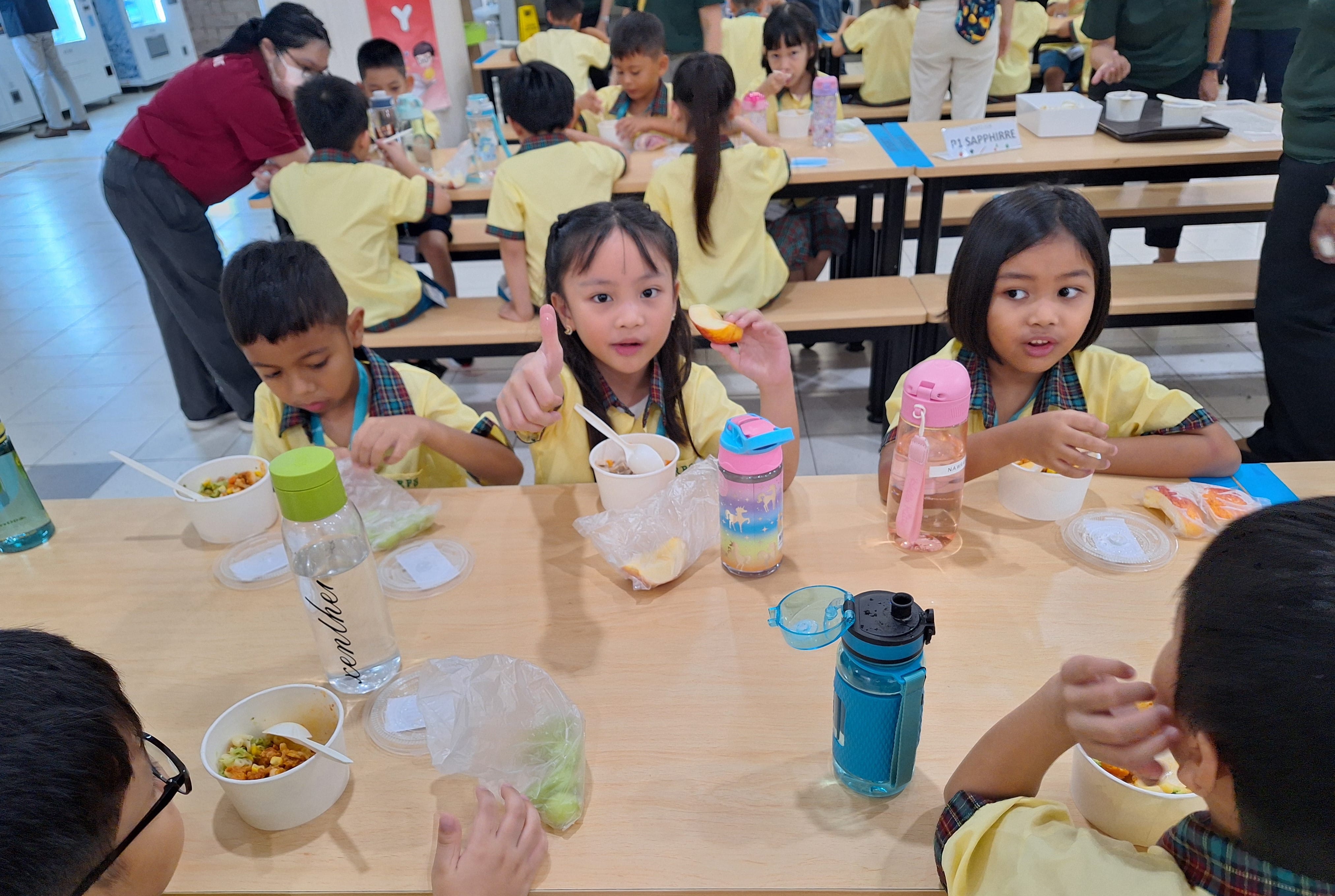 Young children in yellow uniforms eating lunch at long wooden tables in a school cafeteria.