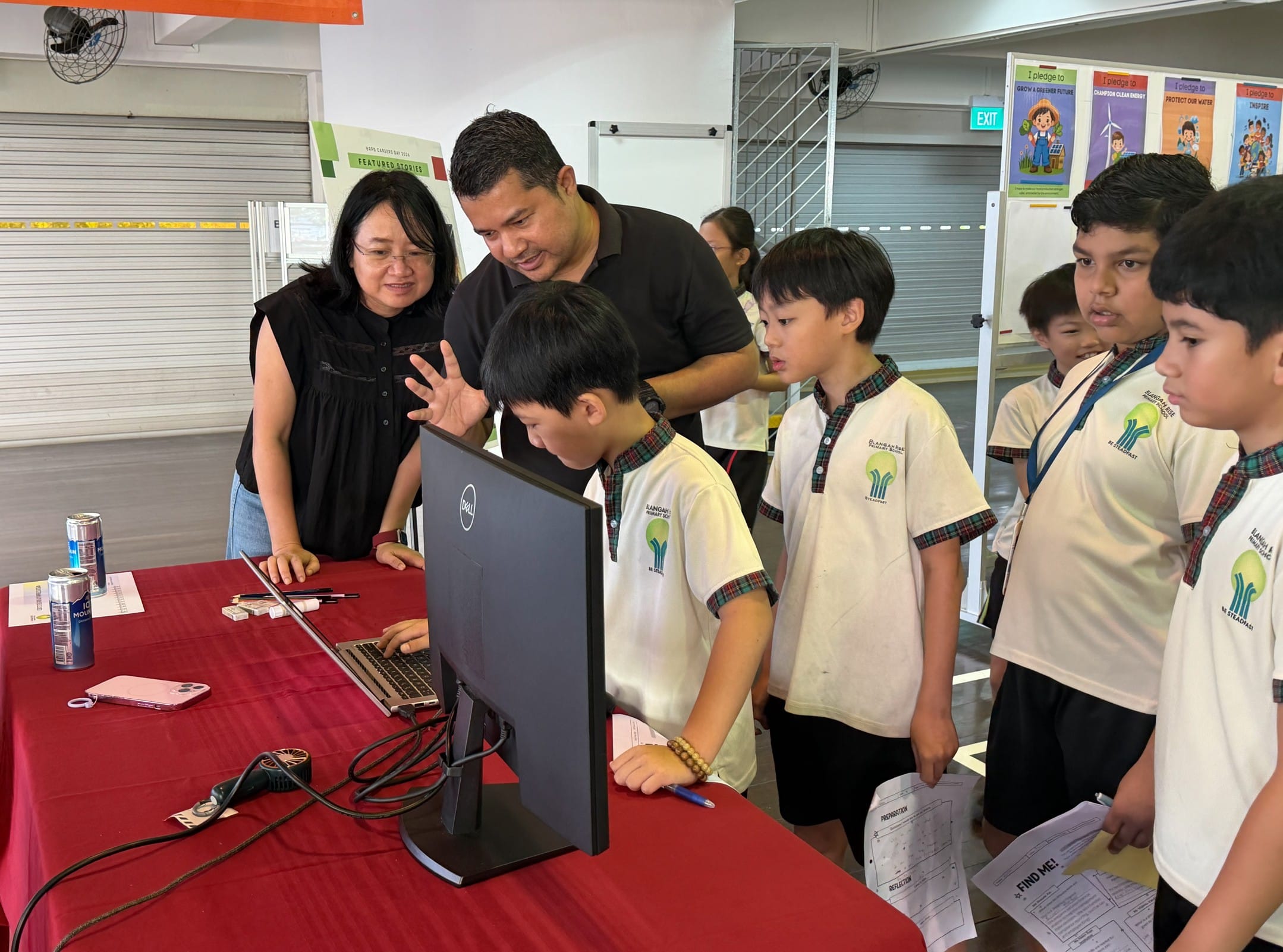 Two adults guide uniformed primary school students gathered around a Dell monitor and laptop on a red table.