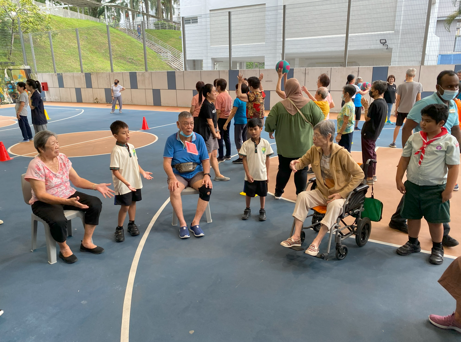 Group of seniors and children on an outdoor court; some seated, one elderly person in a wheelchair.