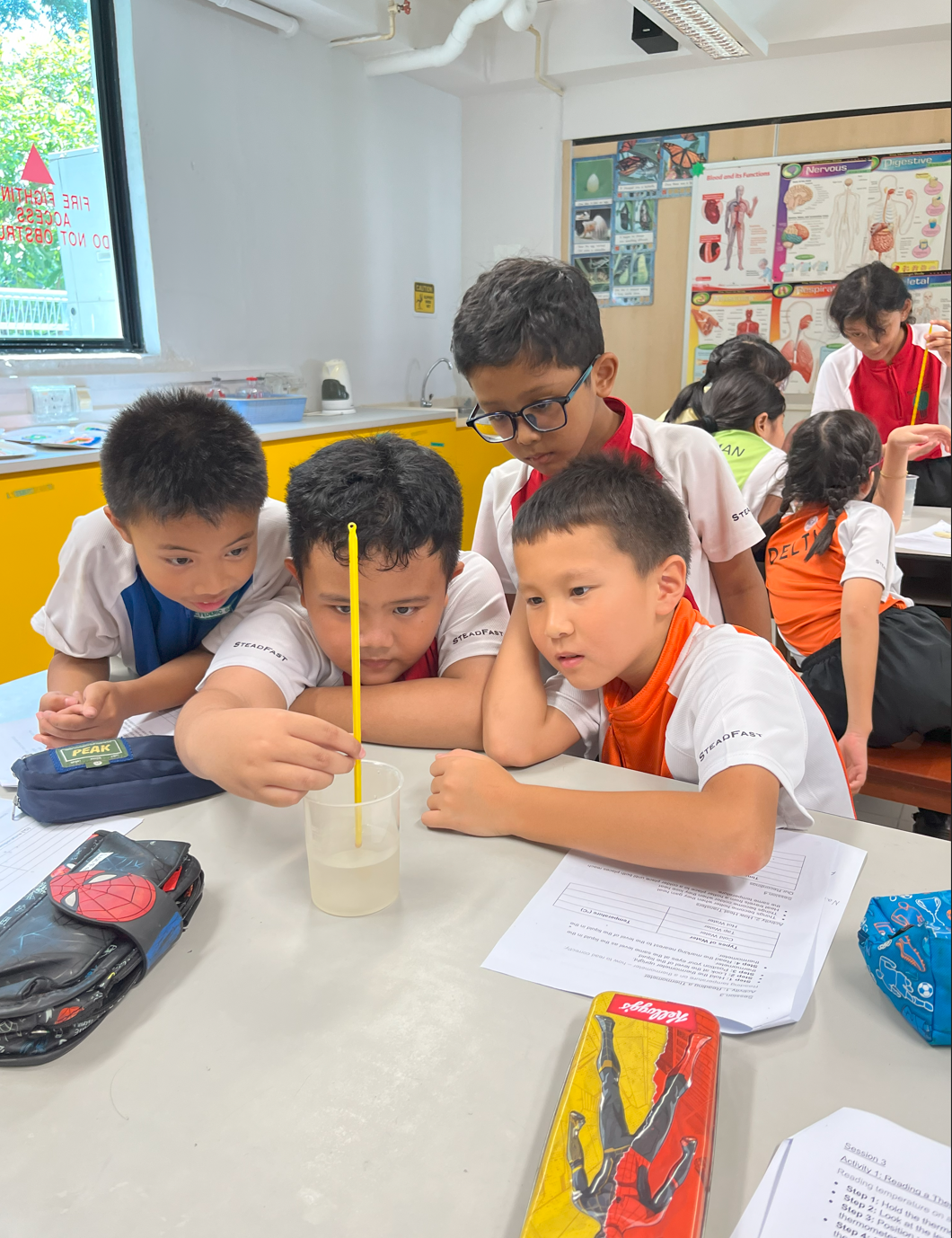 Four young students in school uniforms huddle around a clear cup of liquid with a yellow stirring rod during a science class.