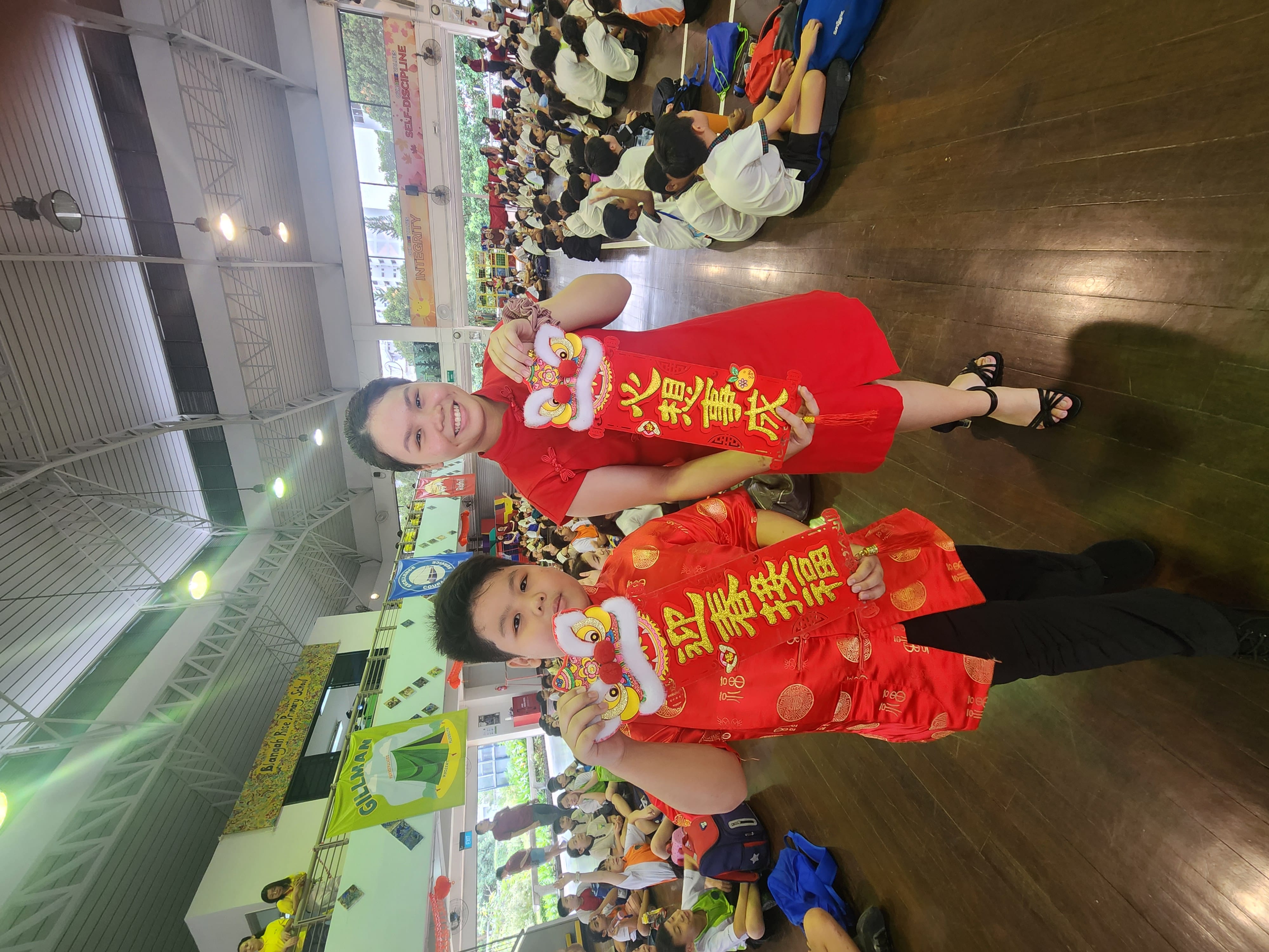 Two children in red traditional Chinese outfits hold lion head decorations and red banners at a school Lunar New Year event.