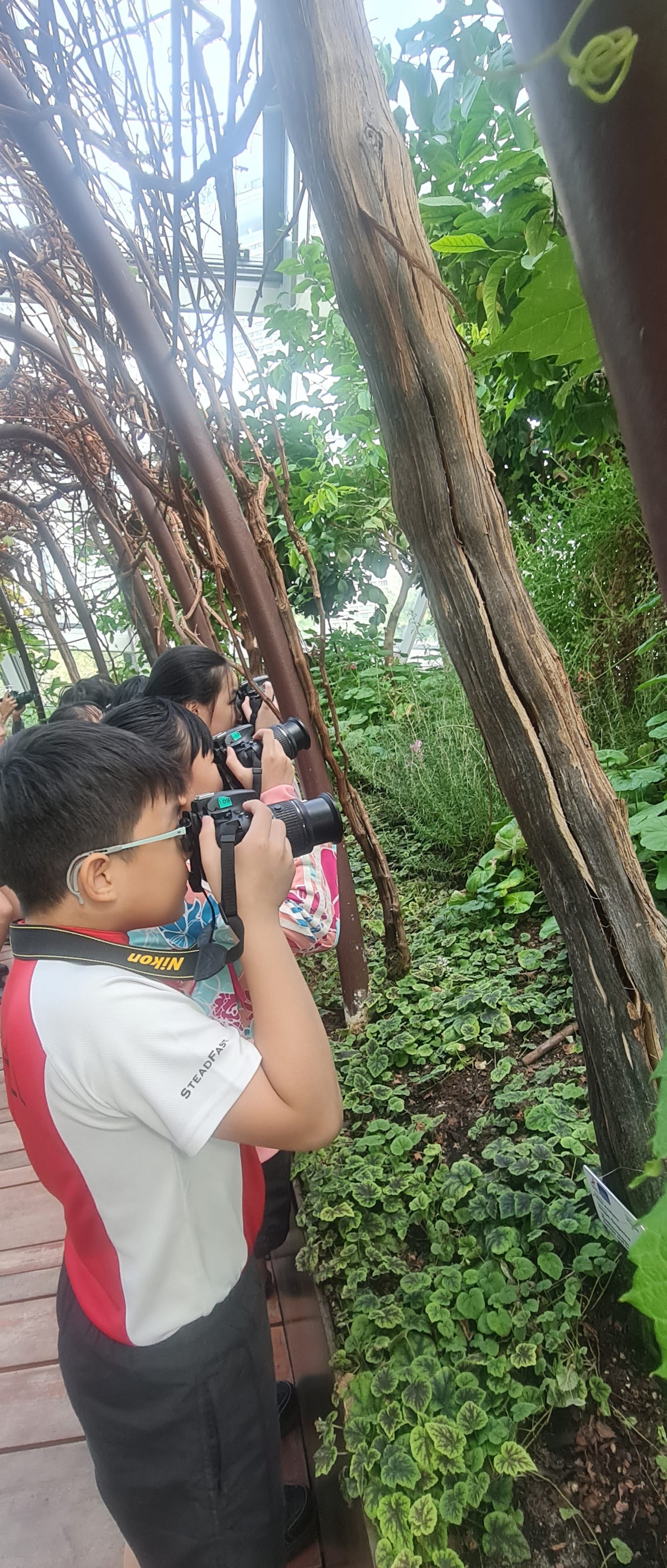 A young boy wearing glasses holds a Nikon camera, photographing plants in a lush greenhouse alongside other children.