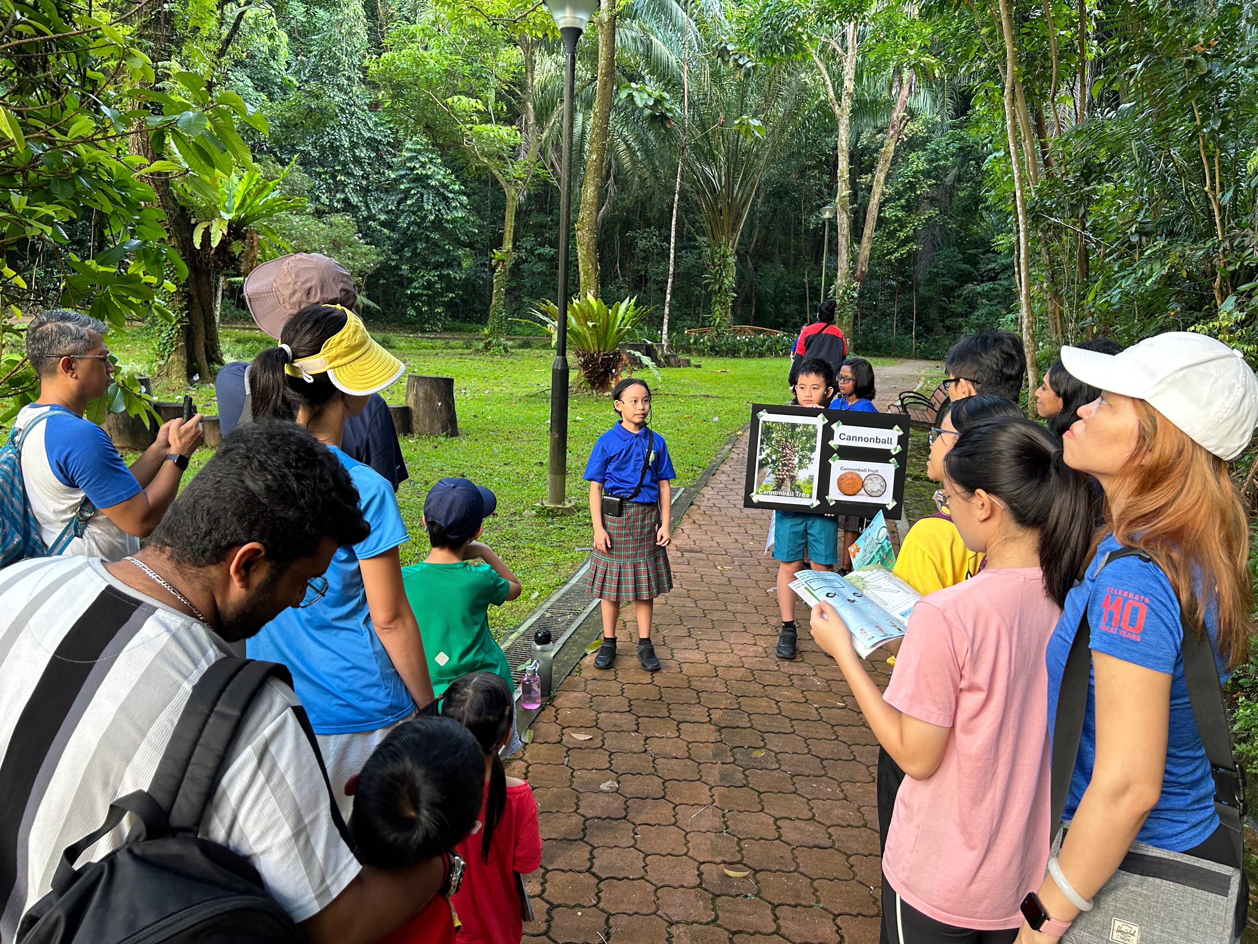 Group on a forest path watches children presenting a poster about cannonball fruit.