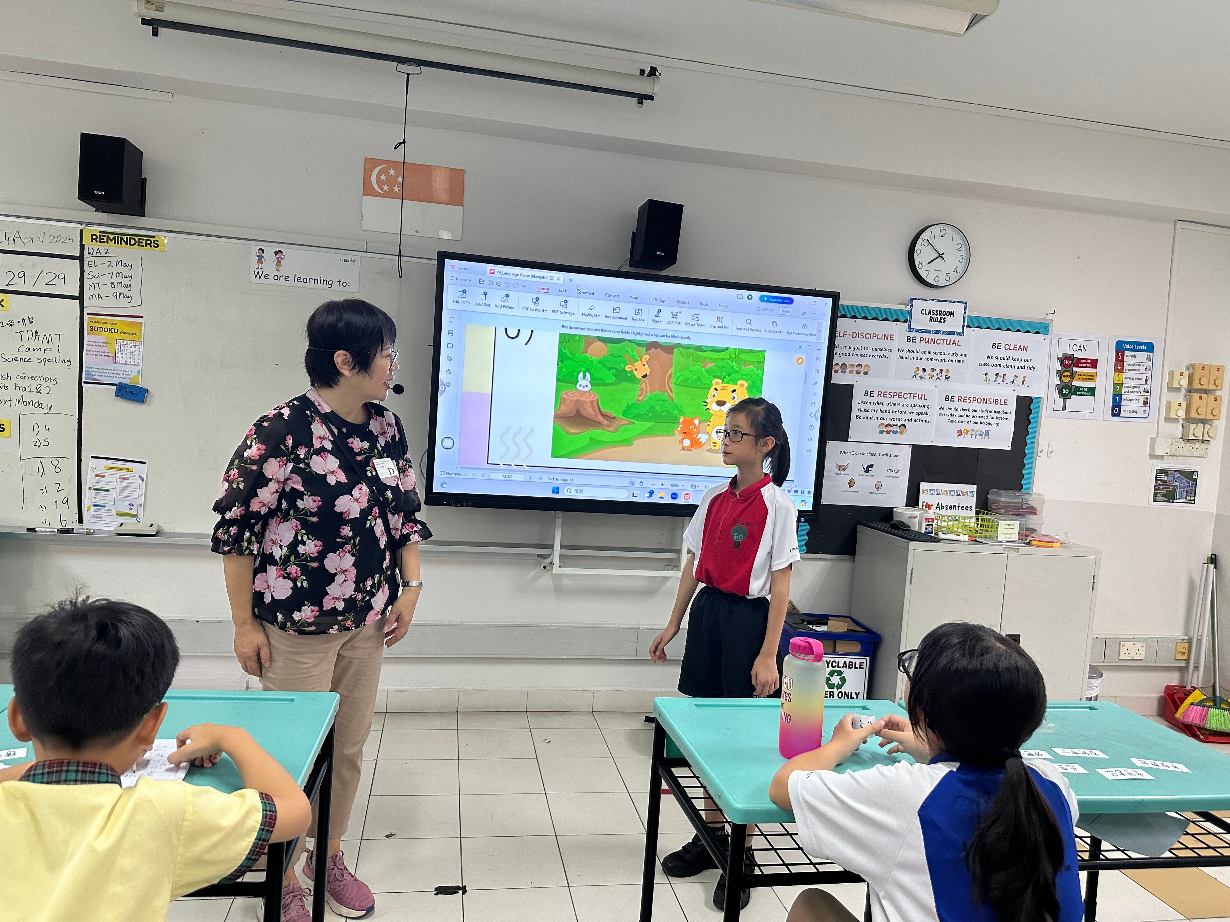 A teacher stands beside a student near a screen showing a cartoon forest scene in a primary school classroom.