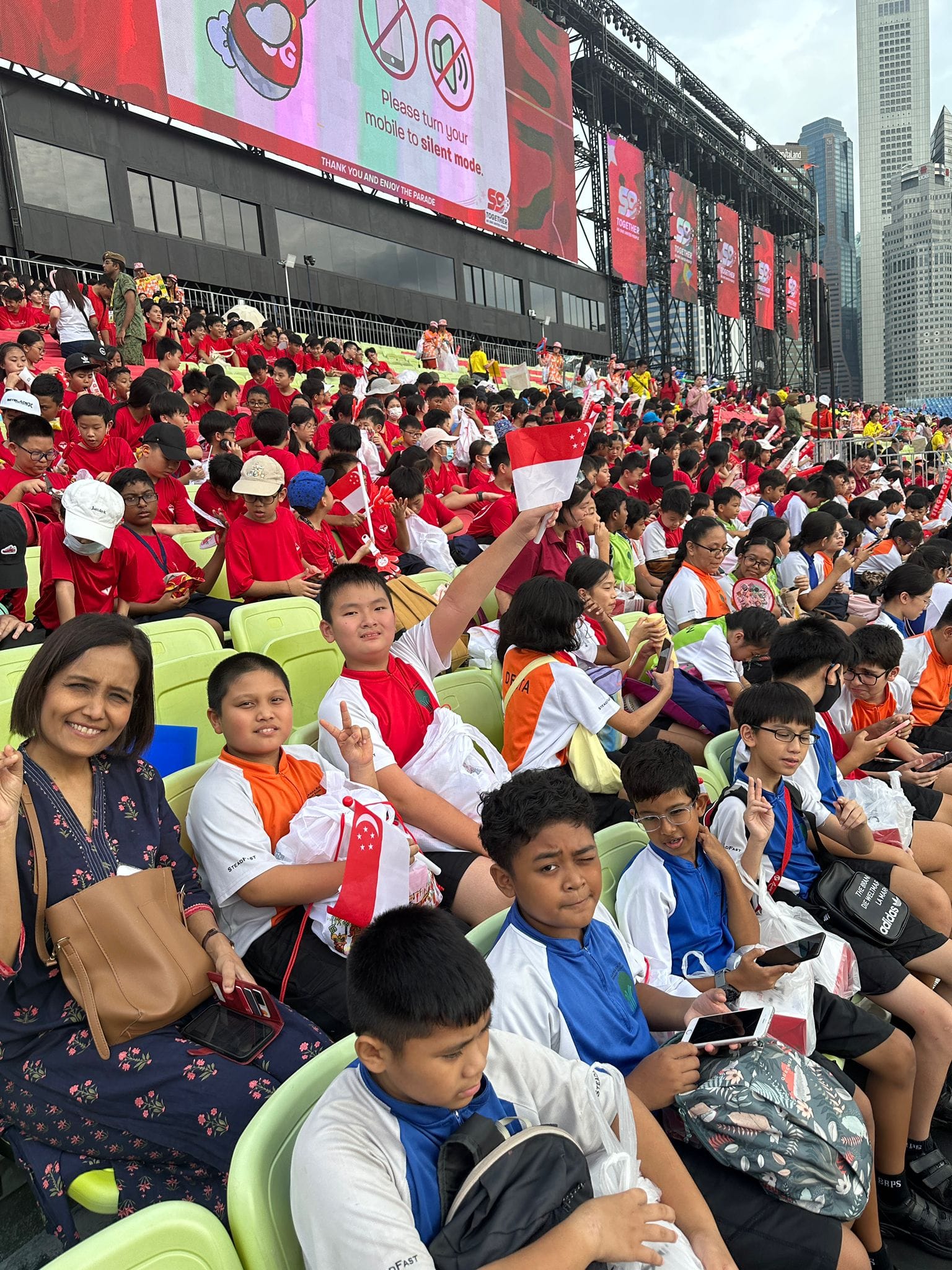 Large crowd of children and adults in red and white outfits seated at Singapore's National Day Parade, waving flags.