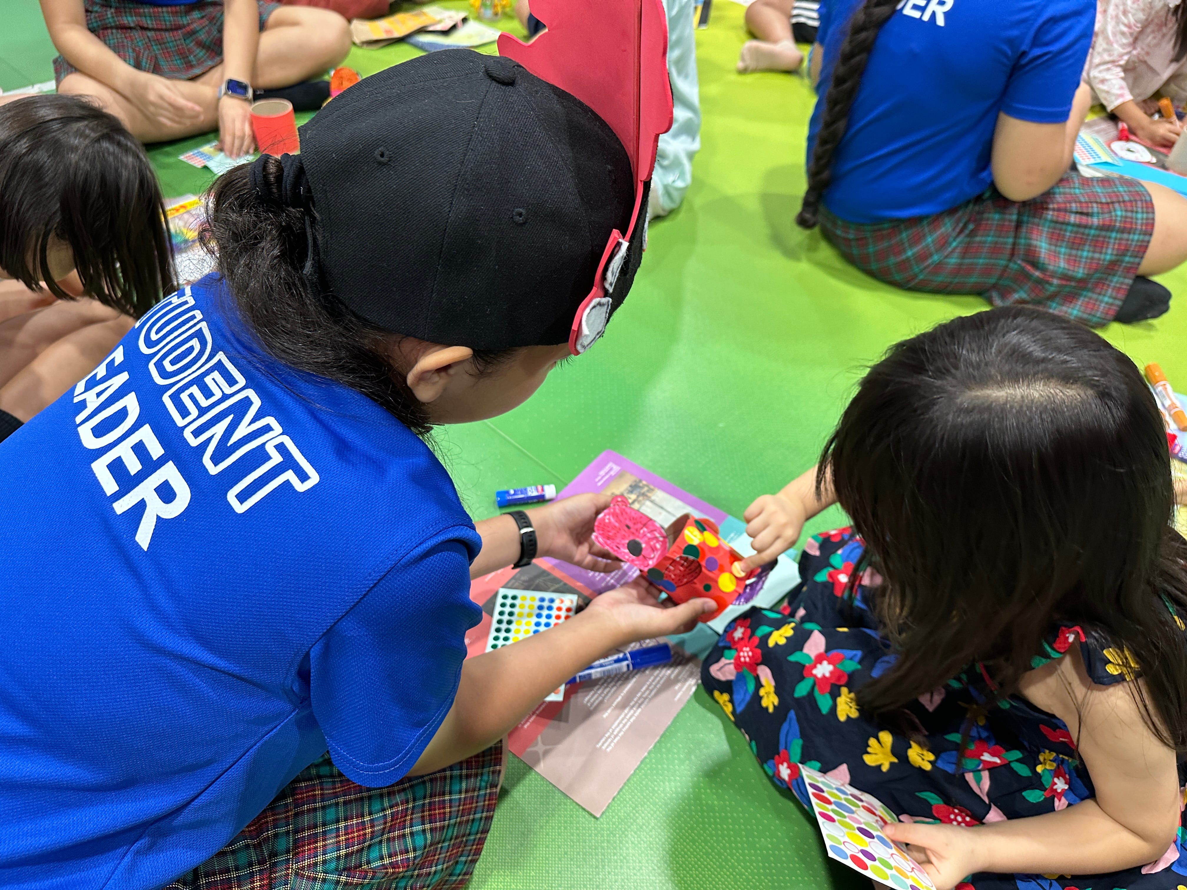 Adult in blue "Student Leader" shirt helps a child place colorful stickers on a paper craft on the floor.