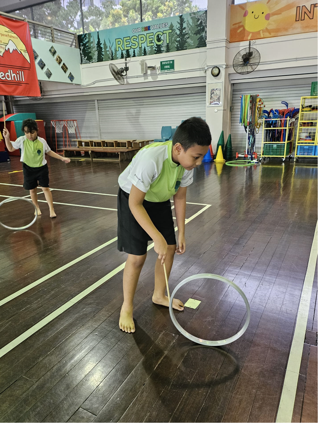Two children in school uniforms play with hoops using sticks on a wooden gym floor indoors.