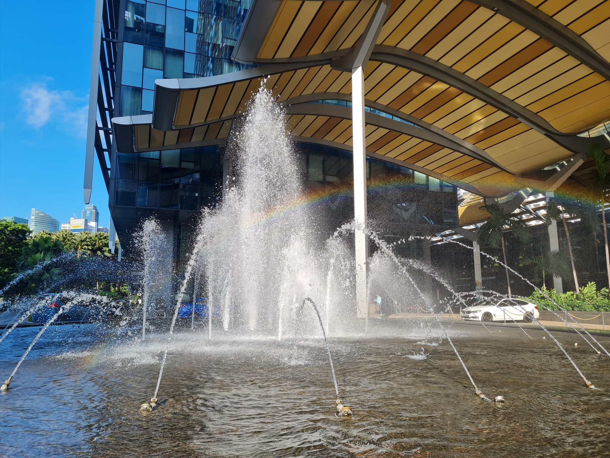 Outdoor water fountain spraying jets near a modern glass building with a rainbow visible in the mist.