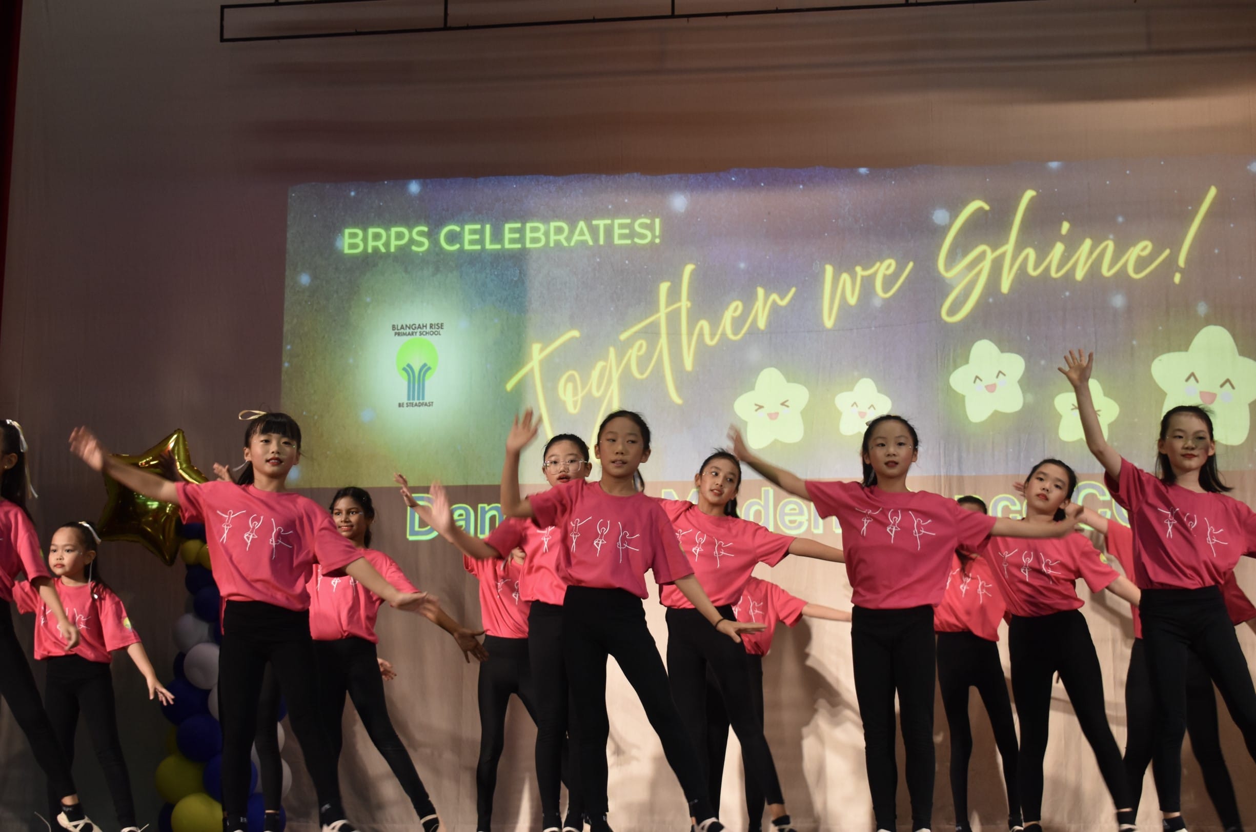 Girls in pink shirts and black pants perform a dance on stage at a BRPS Celebrates Together We Shine school event.