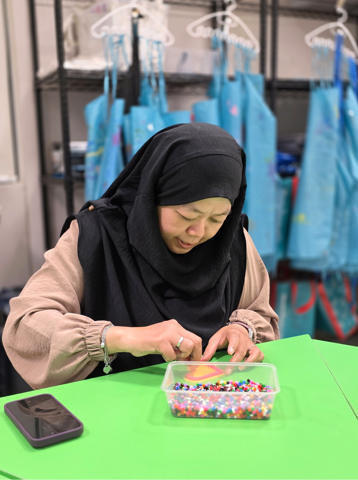 Woman in black hijab sorting colorful beads from a plastic container at a green table, with blue aprons hanging behind her.