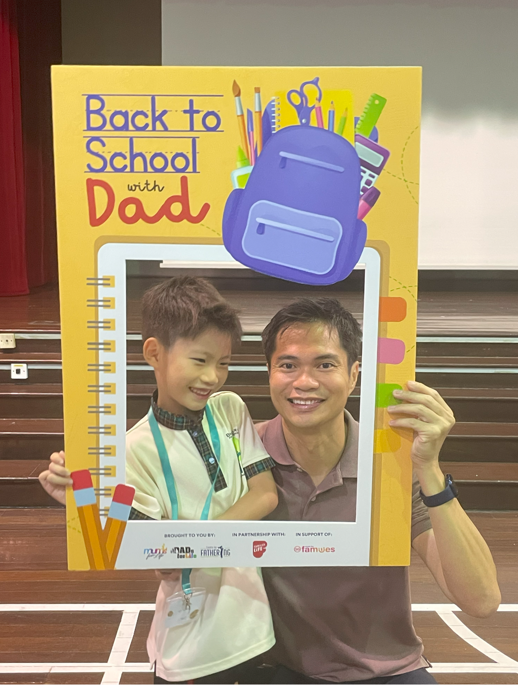 A man and young boy smile through a yellow photo board reading "Back to School with Dad" in a school hall.