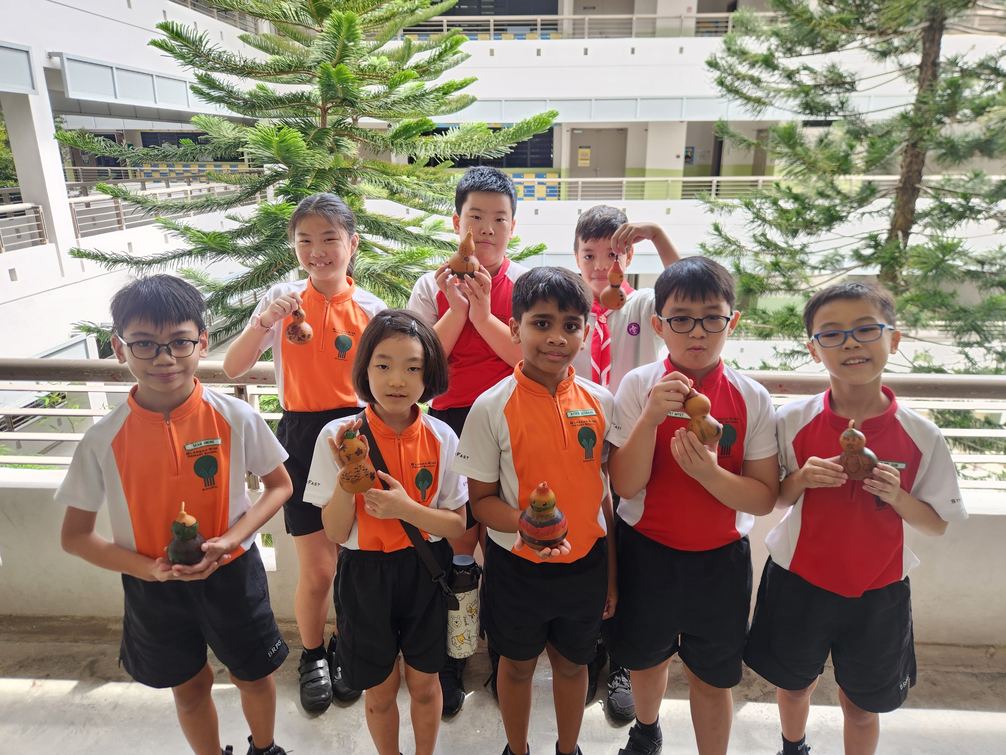 Eight school children in orange and white uniforms stand on a balcony, each holding small handmade figurines.